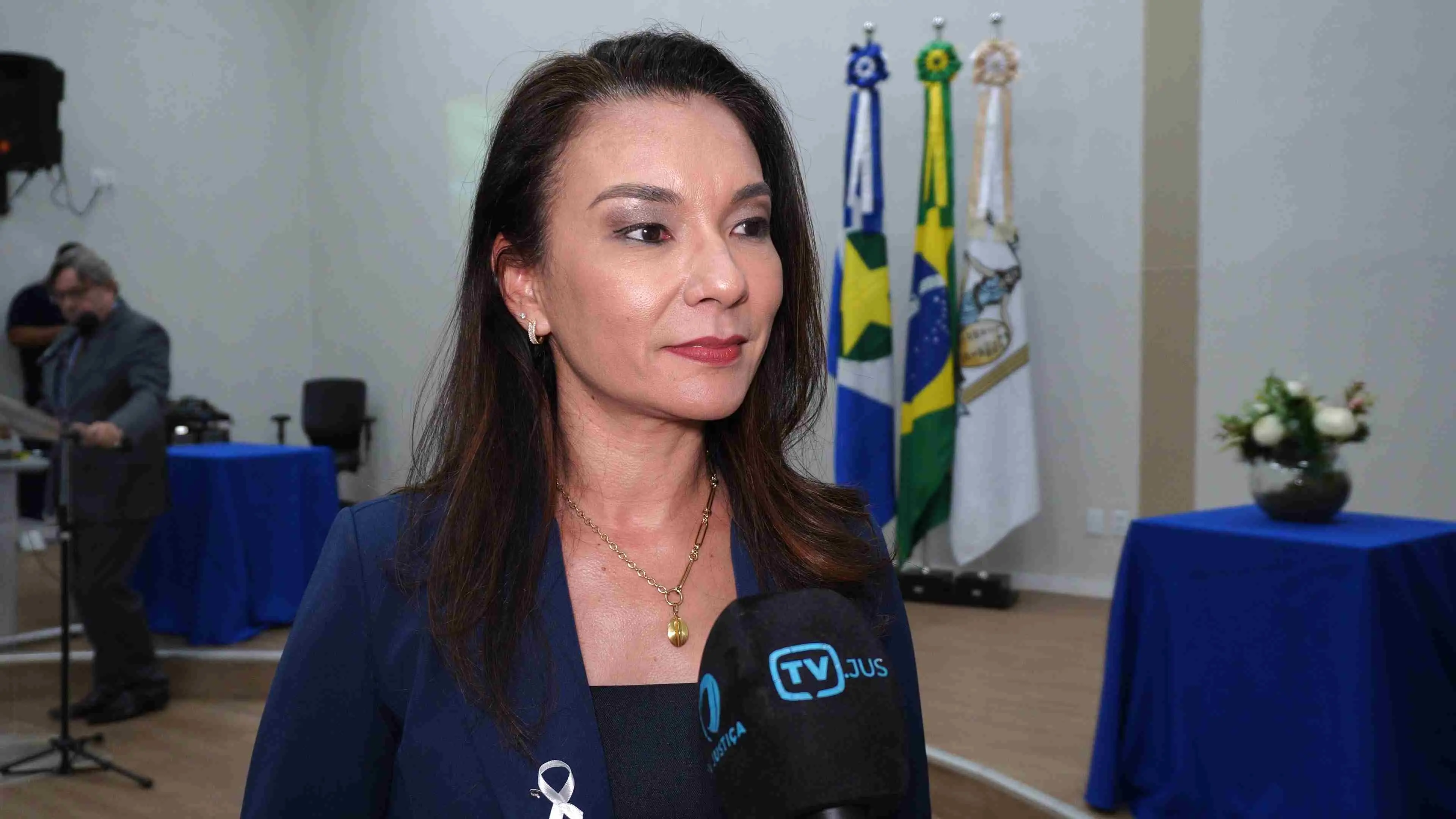 Mulher de blazer azul concede entrevista à TV Justiça. Ao fundo aparecem bandeiras institucionais e mesas cobertas com toalhas azuis, indicando ambiente formal de evento institucional no Fórum de Cuiabá.
