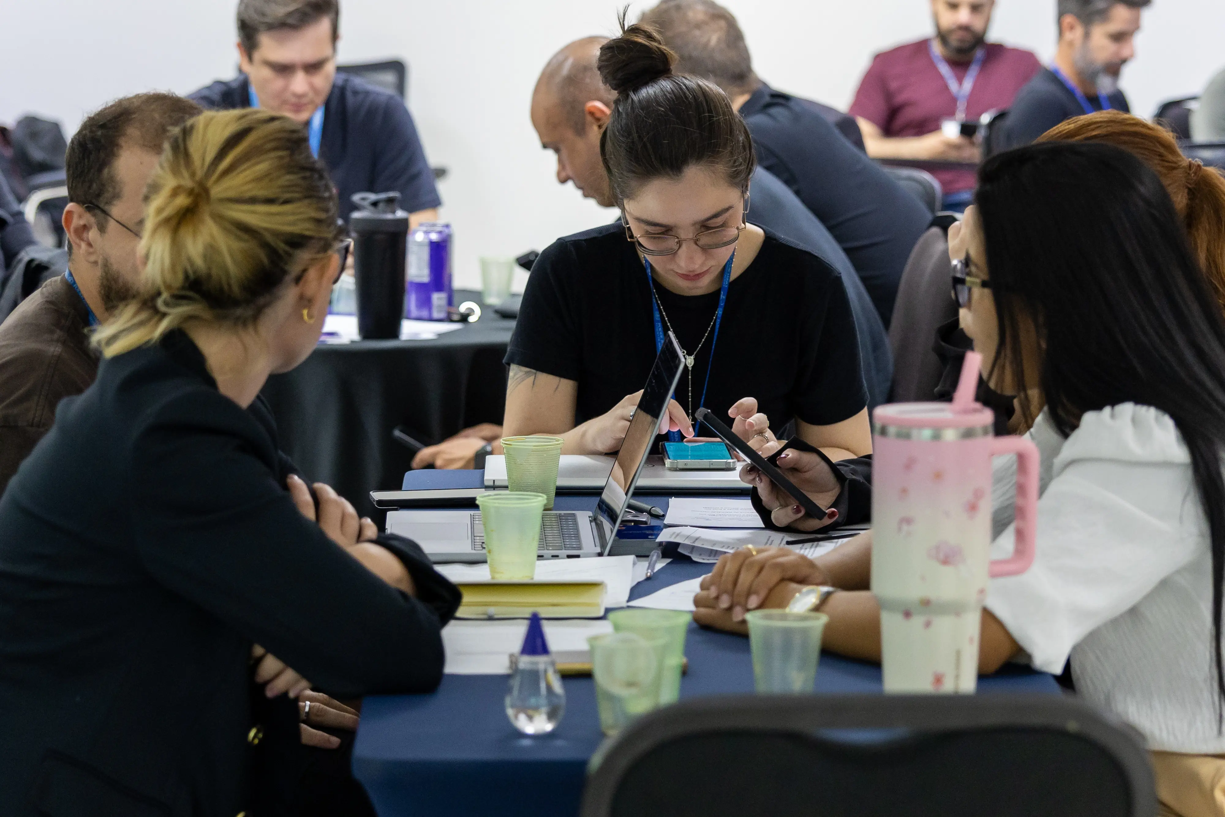 Grupo majoritariamente feminino reunido em torno de mesa azul. Participantes alternam entre celular e notebook, trocando informações e analisando conteúdos impressos durante exercício prático.