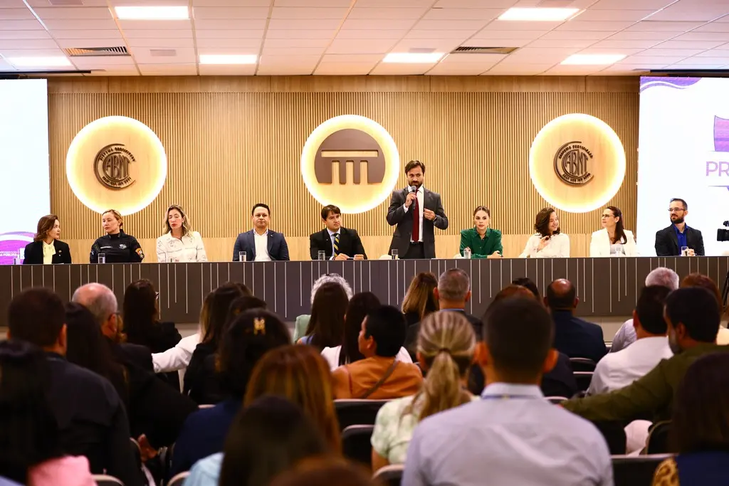 Foto horizontal em plano aberto que mostra o auditório do Tribunal de Contas de Mato Grosso repleto de pessoas assistindo ao juiz Marcelo Resende falar ao microfone no palco, onde há diversas autoridades. 