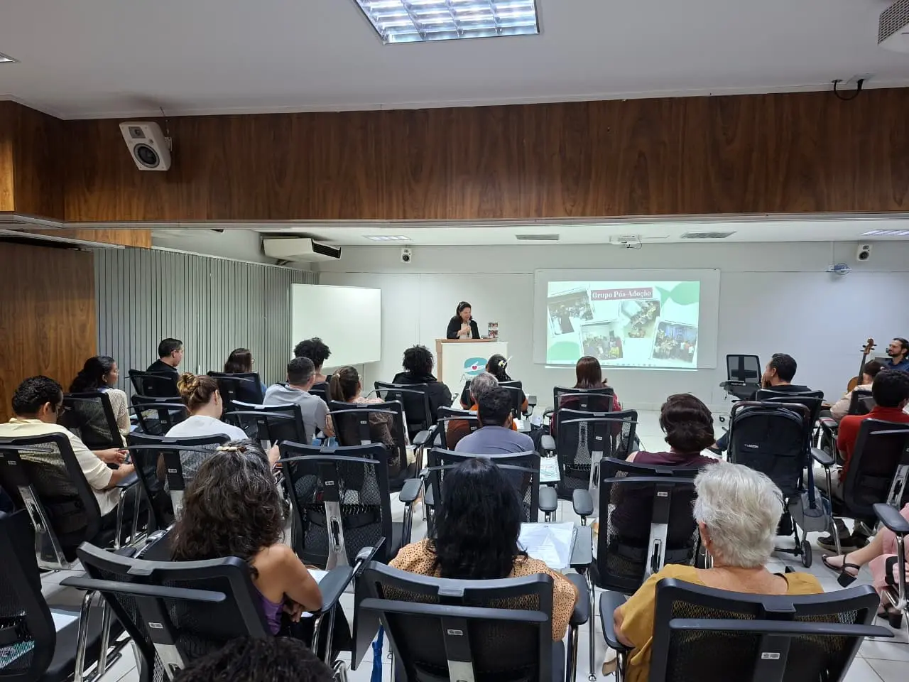 Auditório cheio acompanha apresentação no púlpito do projeto Ampara. Em destaque, slide sobre grupo pós-adoção. Participantes observam atentos, enquanto instrumentos musicais aparecem ao lado do palco.