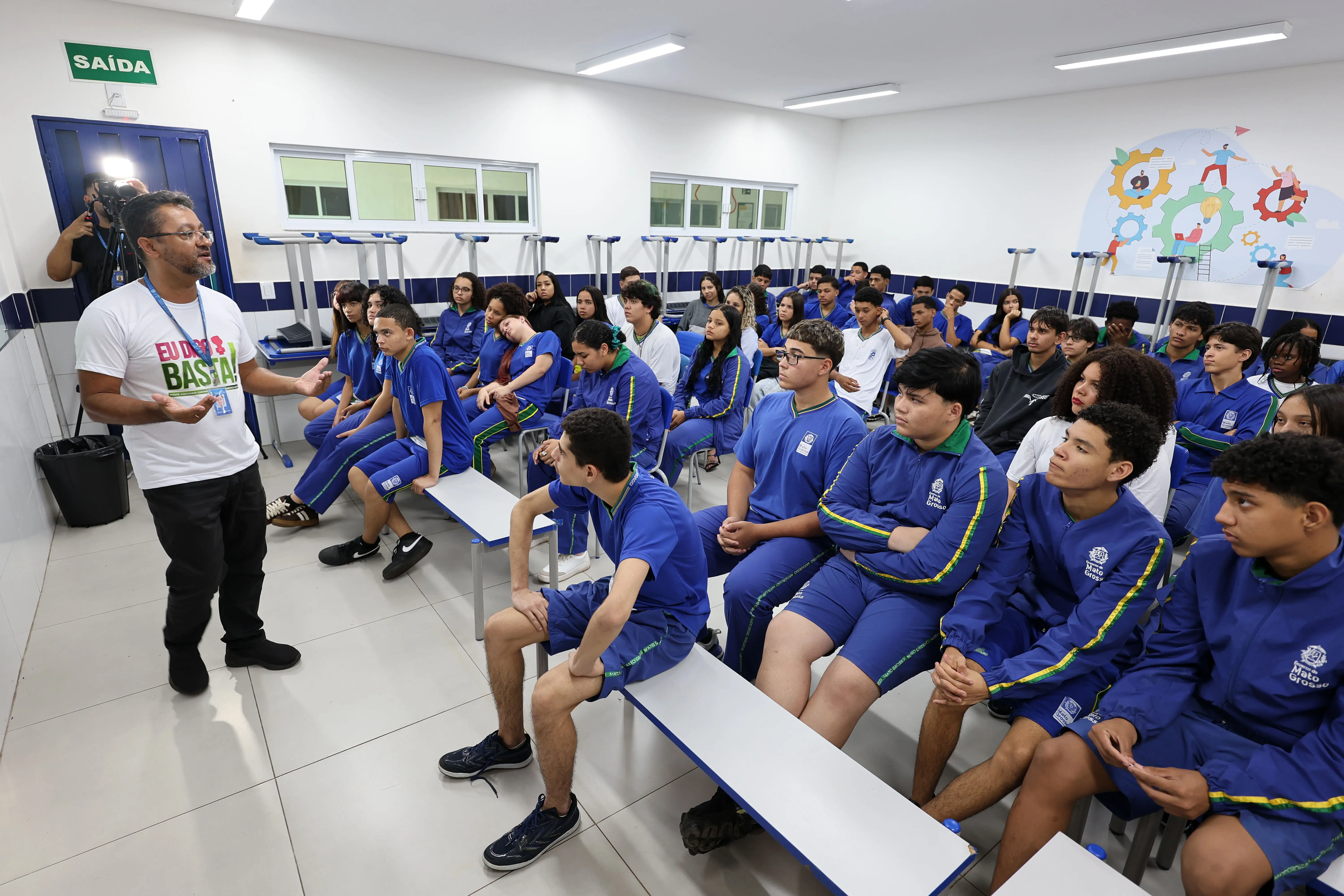 Sala de aula ampla com alunos uniformizados de azul sentados em bancos. À esquerda, o palestrante Cristian Pereira, de camiseta branca com a frase 