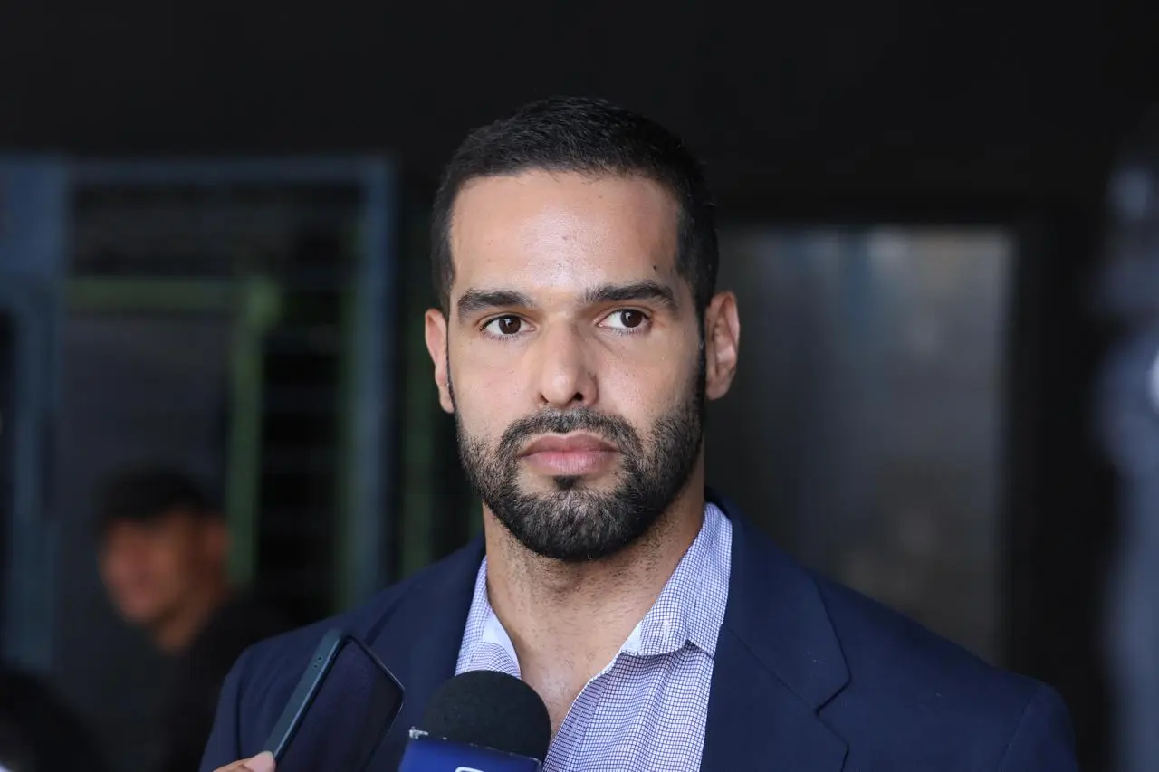 Retrato de um homem de barba e pele clara, vestindo paletó azul e camisa quadriculada. Ele olha para o lado com expressão séria enquanto fala para um microfone e um celular. Fundo escuro.