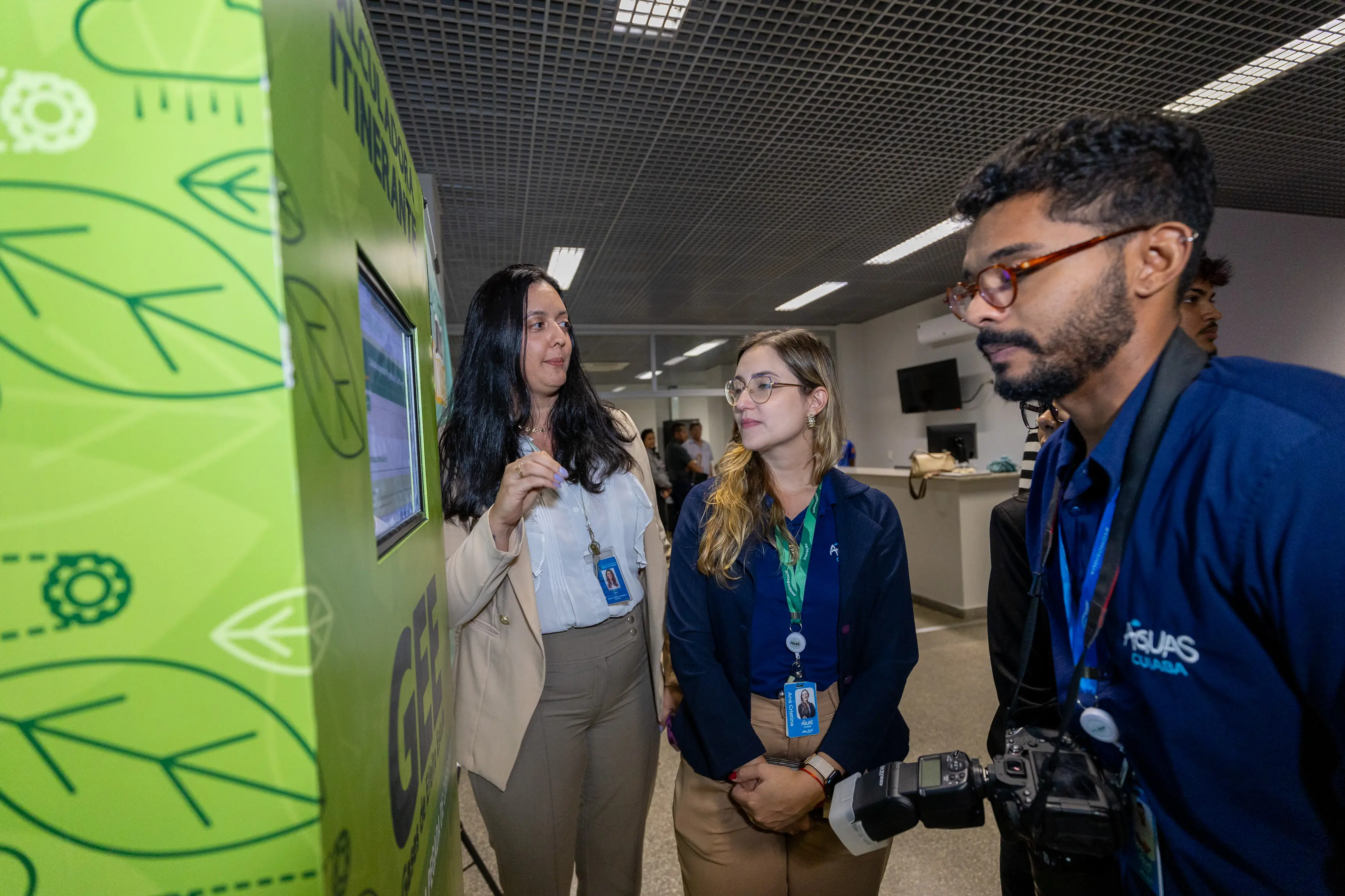  Três pessoas observam um totem verde com ilustrações de folhas e engrenagens. À direita, um homem de barba e óculos segura uma câmera profissional. O ambiente é amplo com teto de grade metálica.