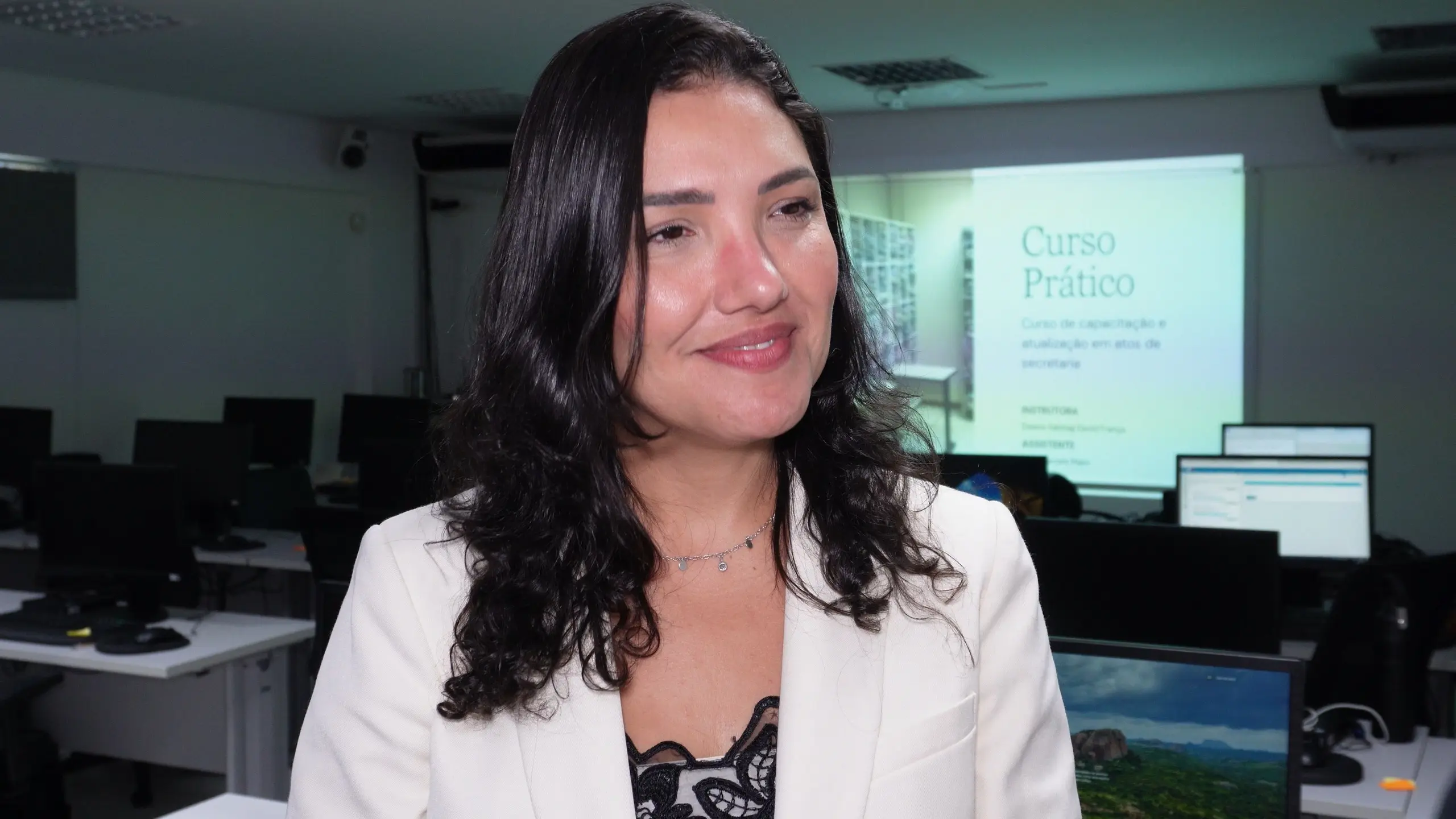 Mulher branca sorri levemente em primeiro plano. Ela tem cabelos escuros ondulados e veste um blazer branco sobre blusa de renda. Ao fundo, uma projeção indica "Curso Prático".