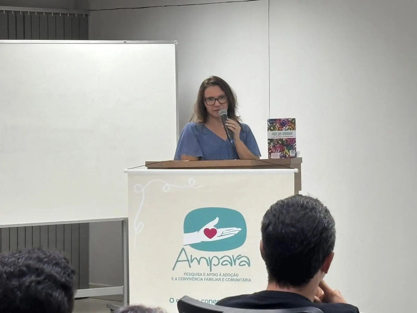Mulher fala ao microfone atrás de um púlpito com a marca Ampara, voltada ao apoio à adoção e convivência familiar. Participantes sentados assistem à apresentação em ambiente de auditório.