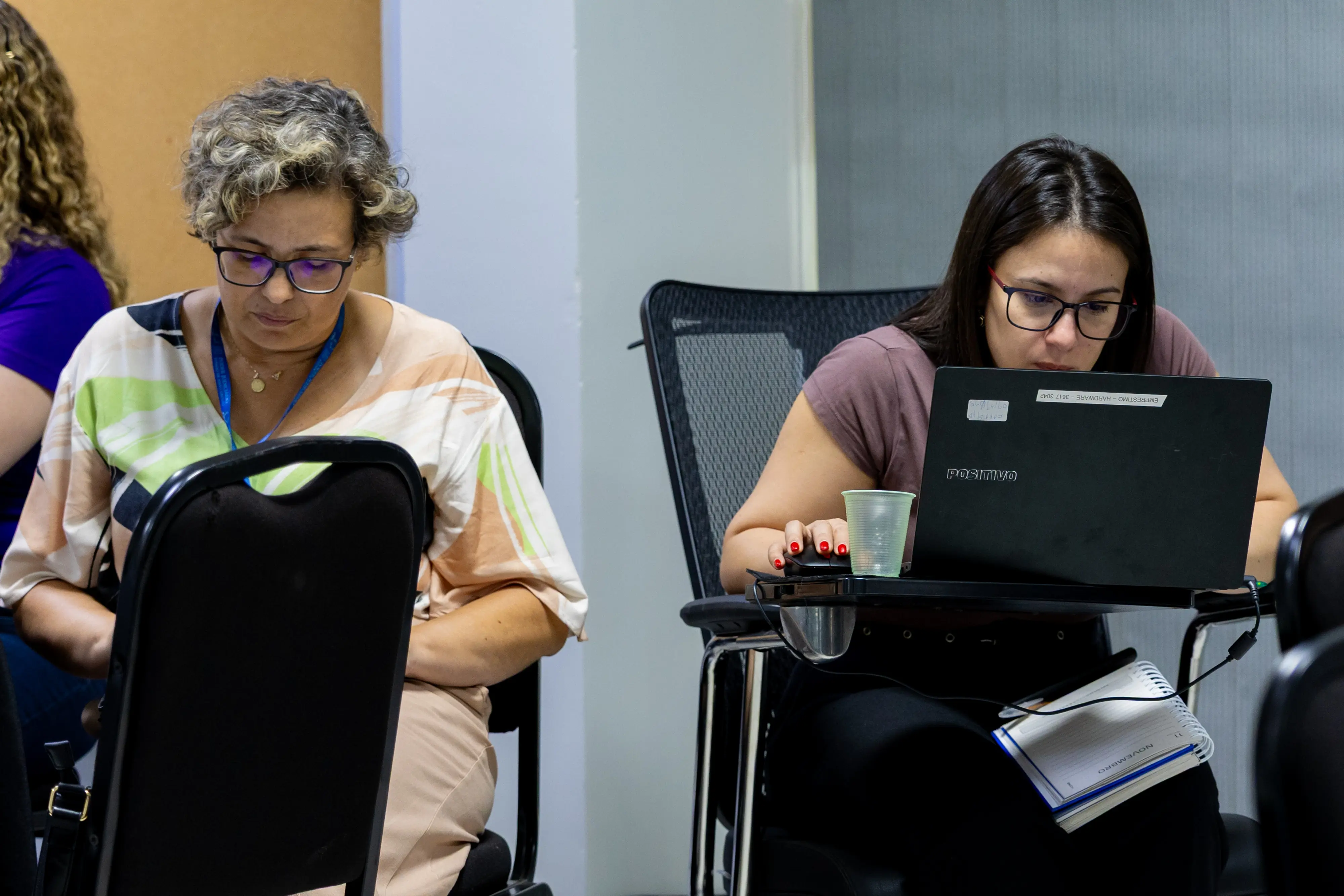 Duas mulheres participam da capacitação. Uma usa notebook apoiado em suporte improvisado; a outra consulta o celular. Ambas demonstram atenção e foco na proposta da oficina.