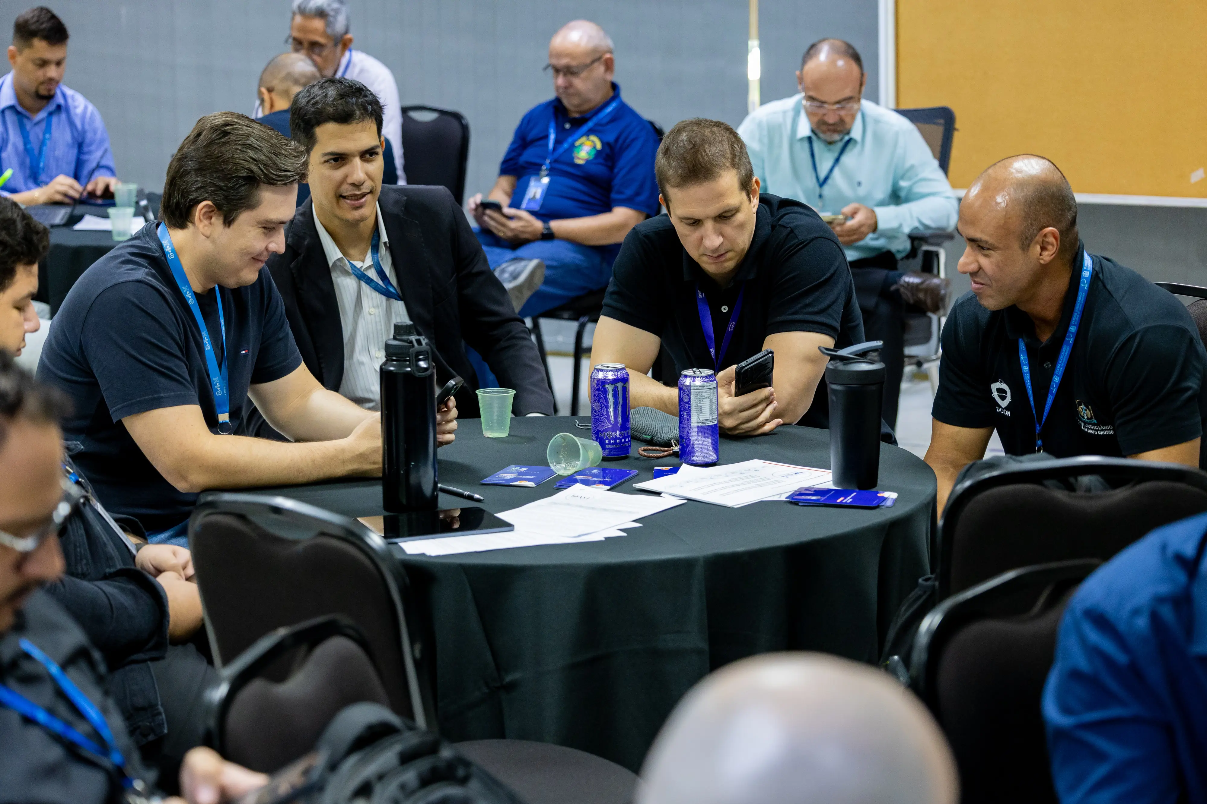 Cinco homens reunidos discutem conteúdo exibido no celular. Garrafas térmicas e latas sobre a mesa acompanham anotações e fichas distribuídas durante a capacitação.