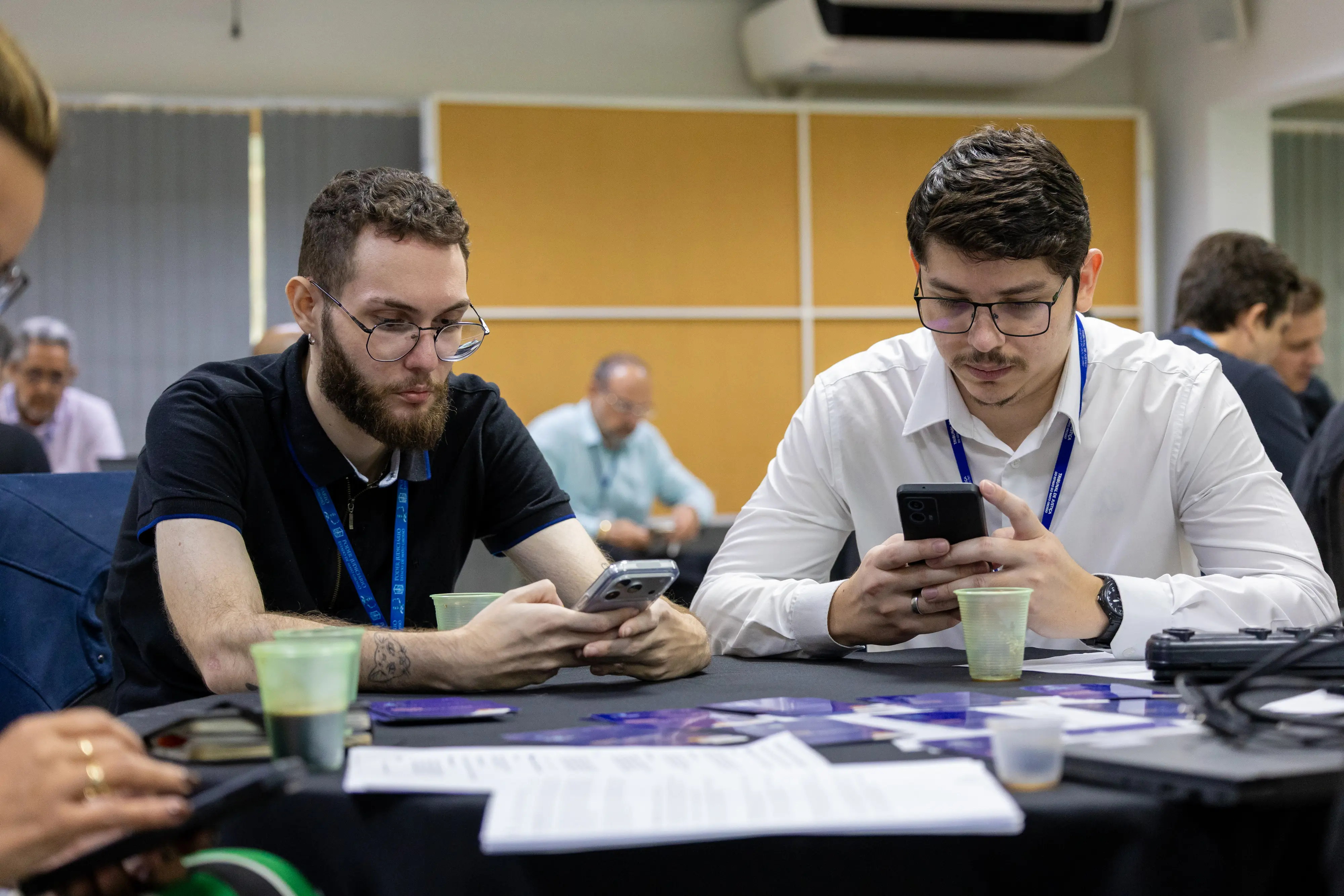 Dois participantes, um de camisa preta e outro branca, concentram-se nos smartphones. À frente, documentos e materiais da oficina espalhados sobre a mesa reforçam o caráter colaborativo.