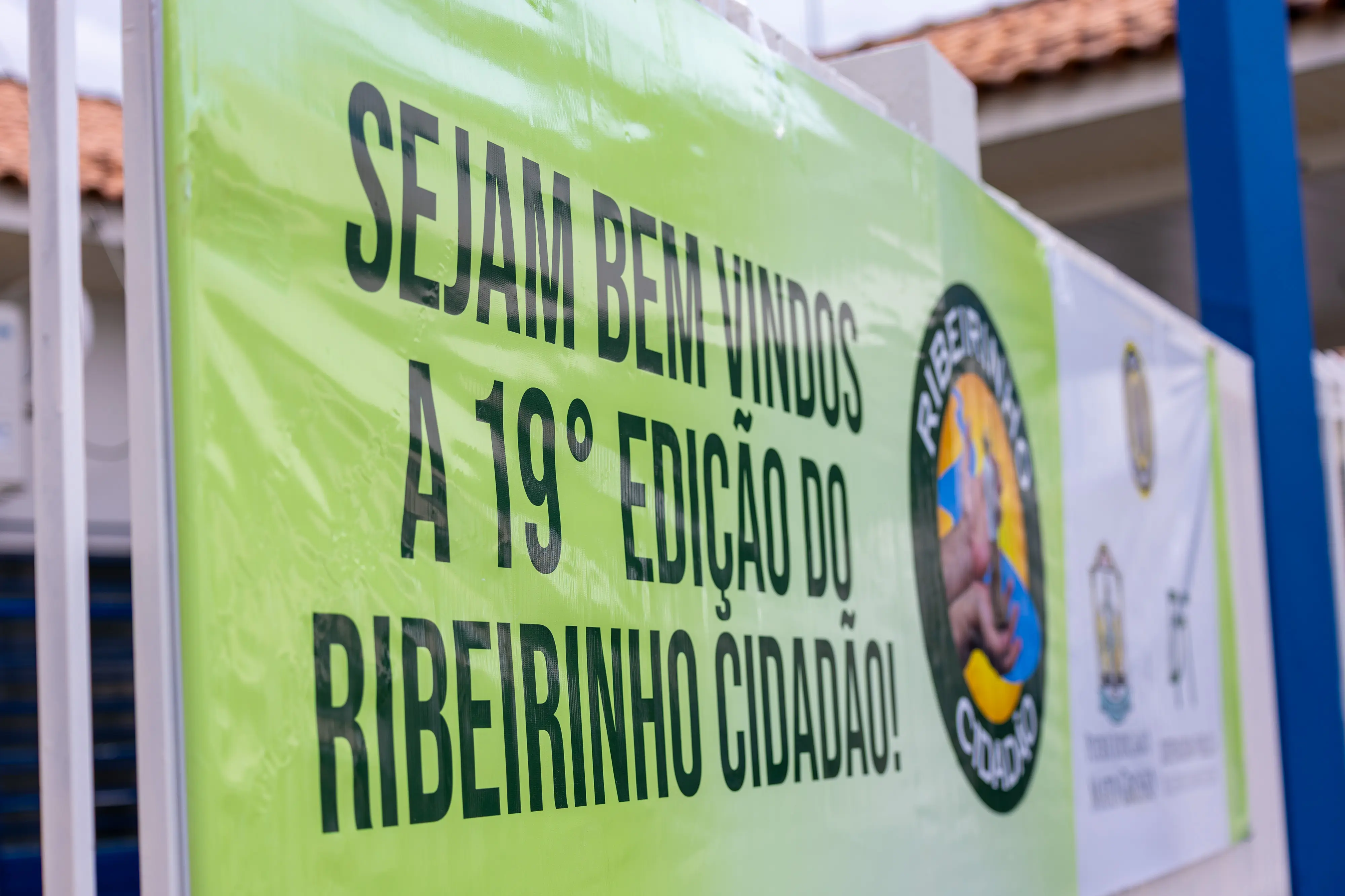 Faixa verde em destaque anuncia “Sejam bem-vindos à 19ª edição do Ribeirinho Cidadão”, com letras pretas em alto contraste. Ao lado, há logotipos institucionais. A placa está fixada em estrutura externa, indicando o local de realização da ação.
