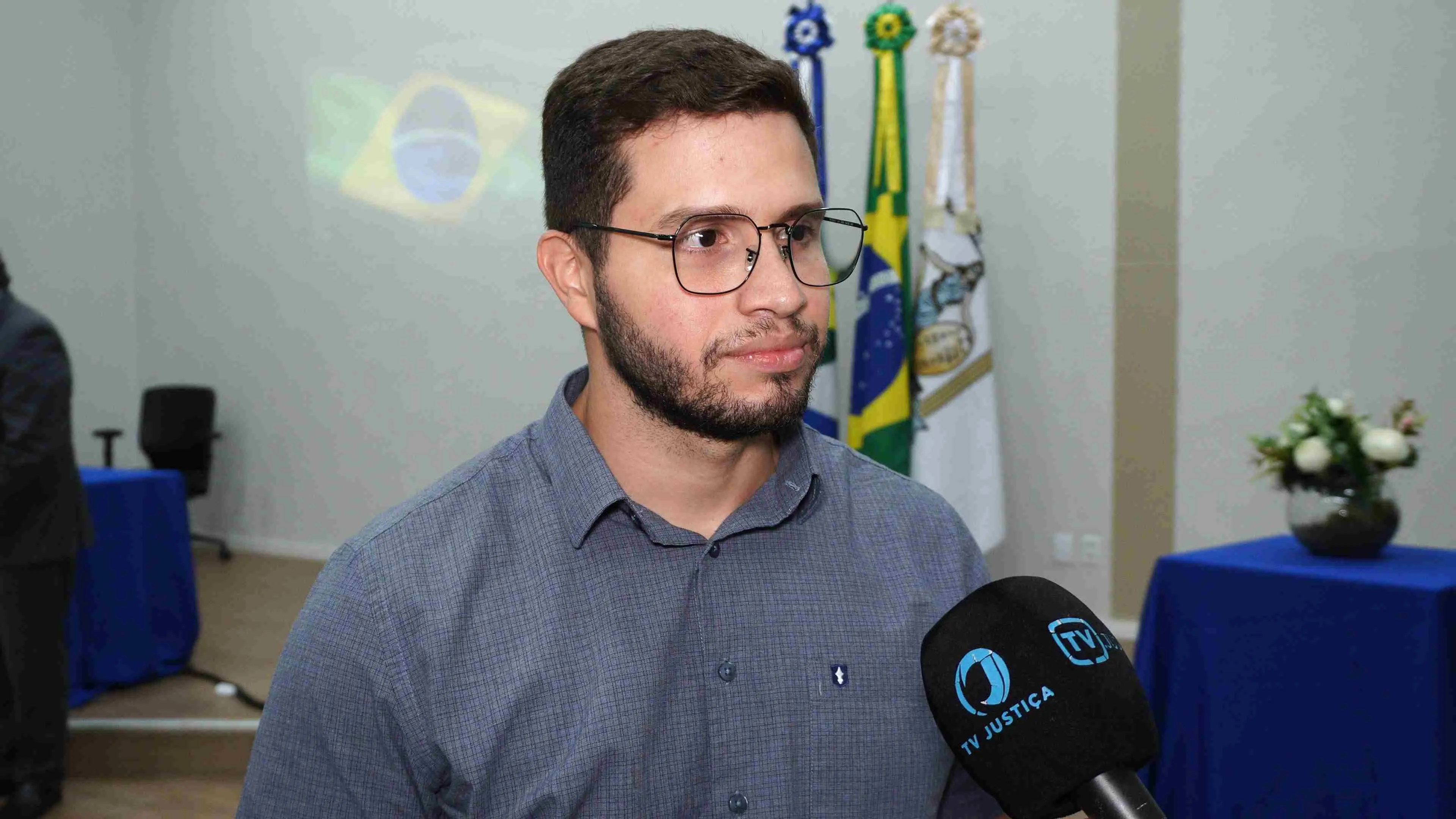 Homem com camisa social cinza e óculos fala à TV Justiça durante entrevista. Ao fundo aparecem bandeiras e parte do ambiente do fórum preparado para a cerimônia de recepção de novos servidores.