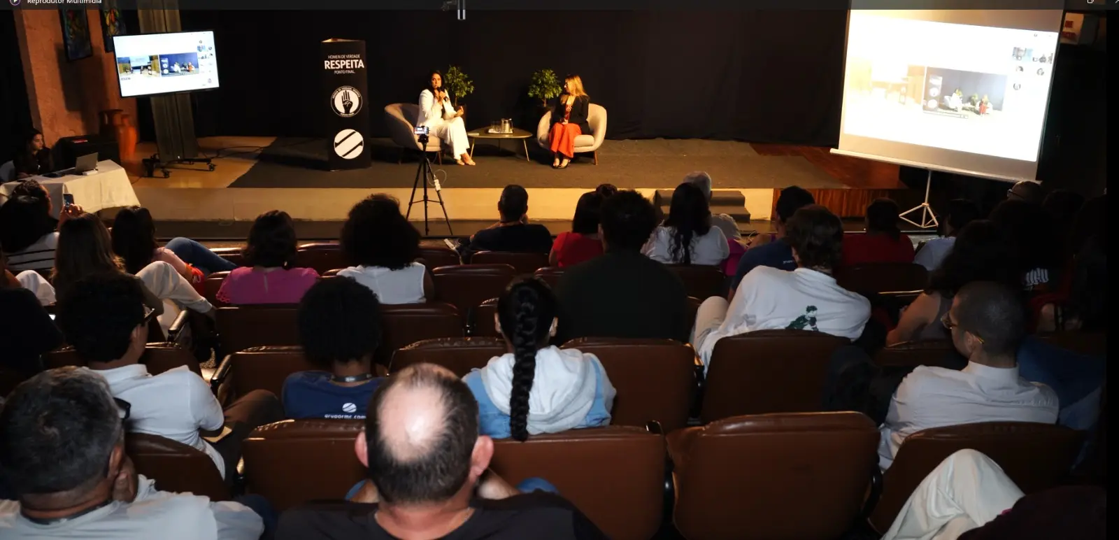 Plateia em poltronas marrons assiste a duas mulheres conversando no palco. Há um totem preto e telões laterais exibindo o evento.