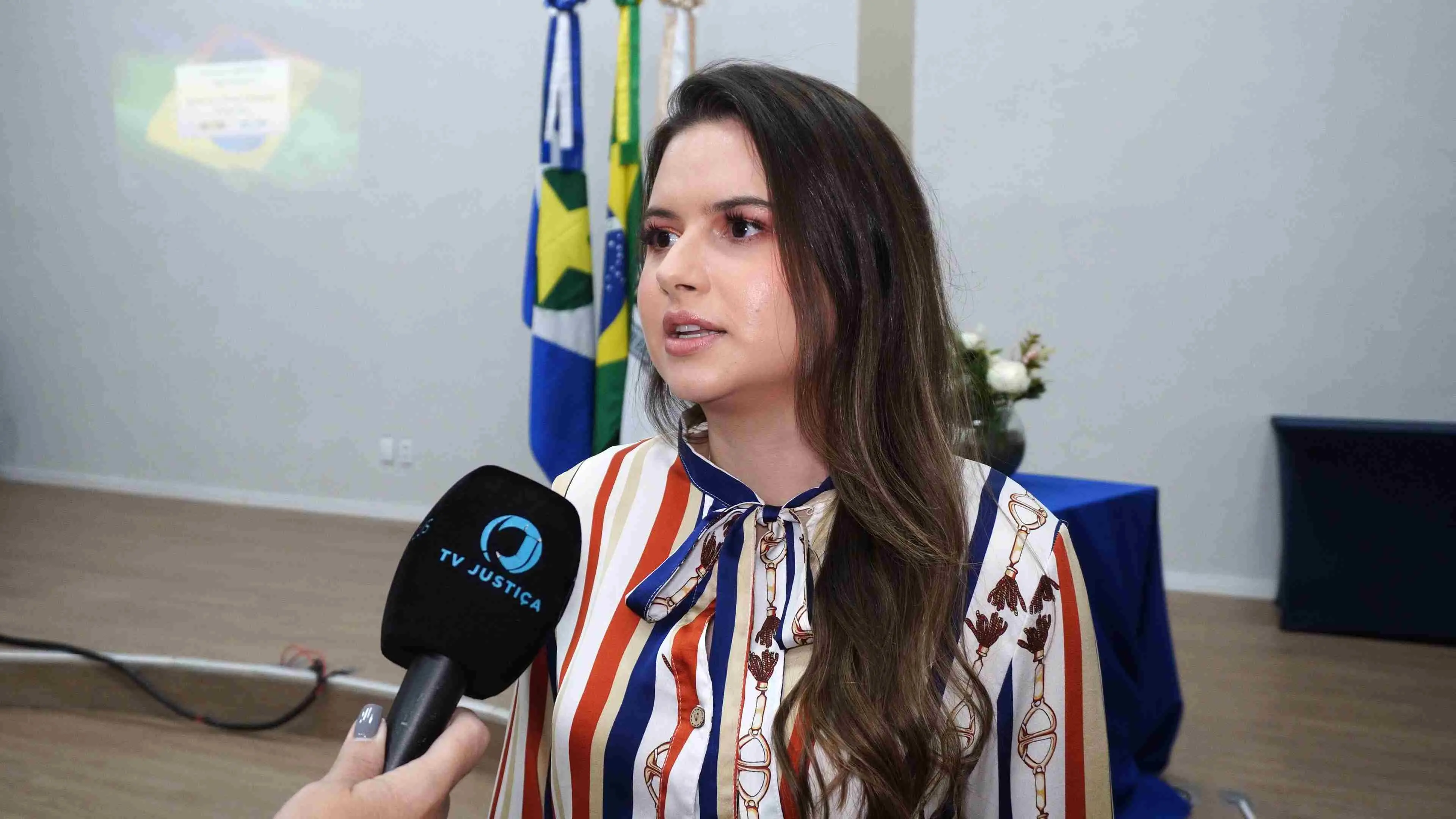 Jovem fala à TV Justiça durante entrevista. Ela veste blusa listrada em tons de azul, vermelho e branco com laço no pescoço. Ao fundo aparecem bandeiras e estrutura do evento realizado no fórum.