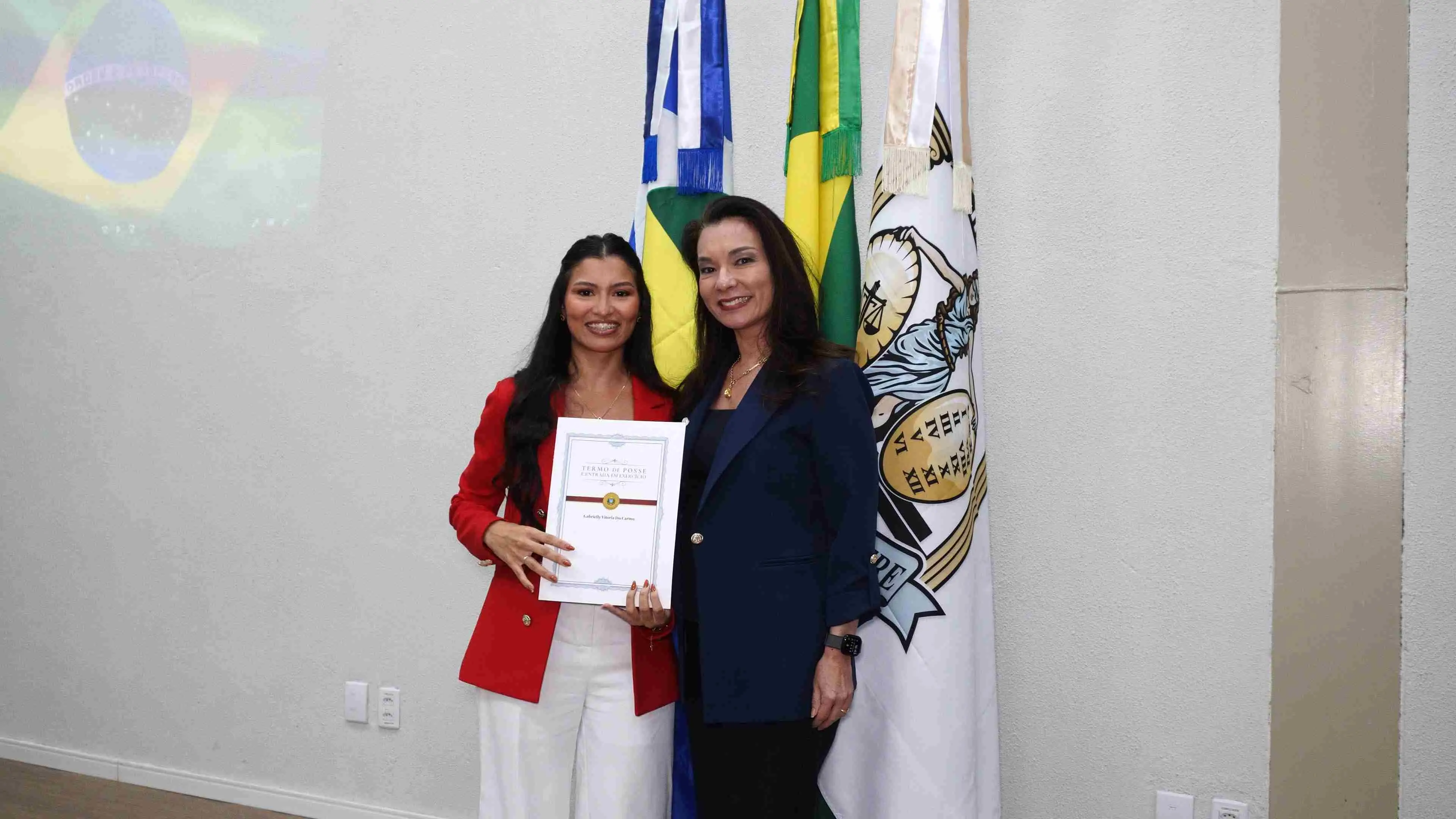 Duas mulheres posam para foto segurando certificado de posse. Uma delas veste blazer vermelho e segura o documento, enquanto a outra usa blazer azul. Bandeiras institucionais aparecem ao fundo da imagem.
