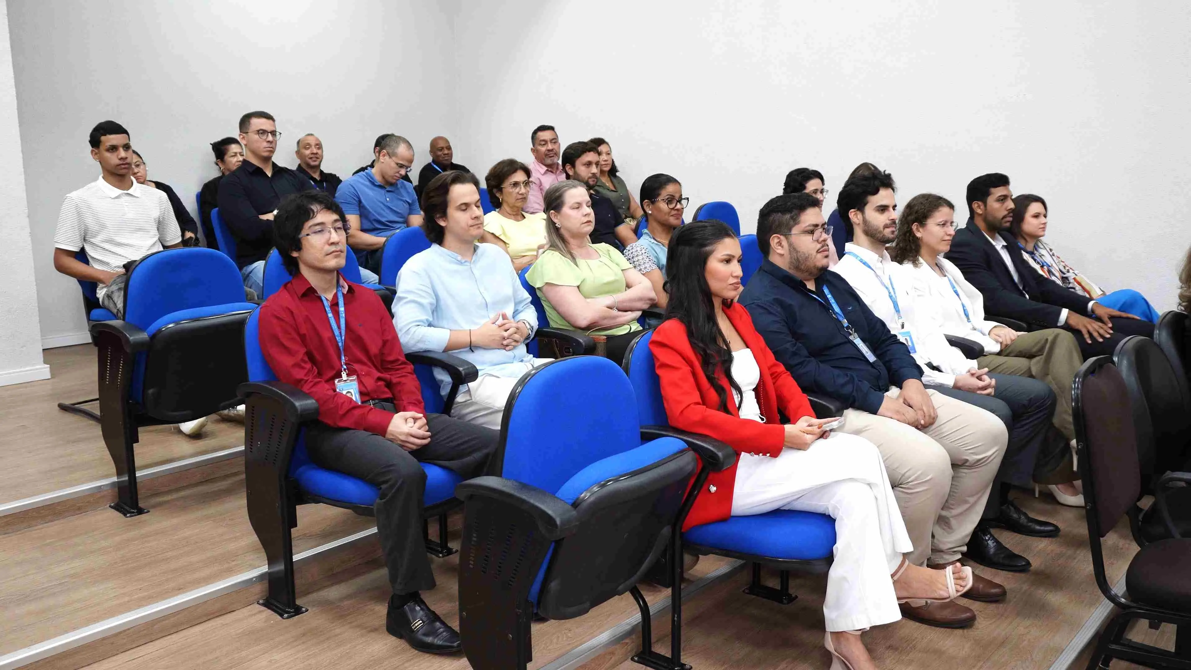 Participantes estão sentados em fileiras de cadeiras azuis em um auditório do fórum. Homens e mulheres, vestidos com trajes formais e crachás institucionais, acompanham atentamente a cerimônia de recepção de novos servidores.