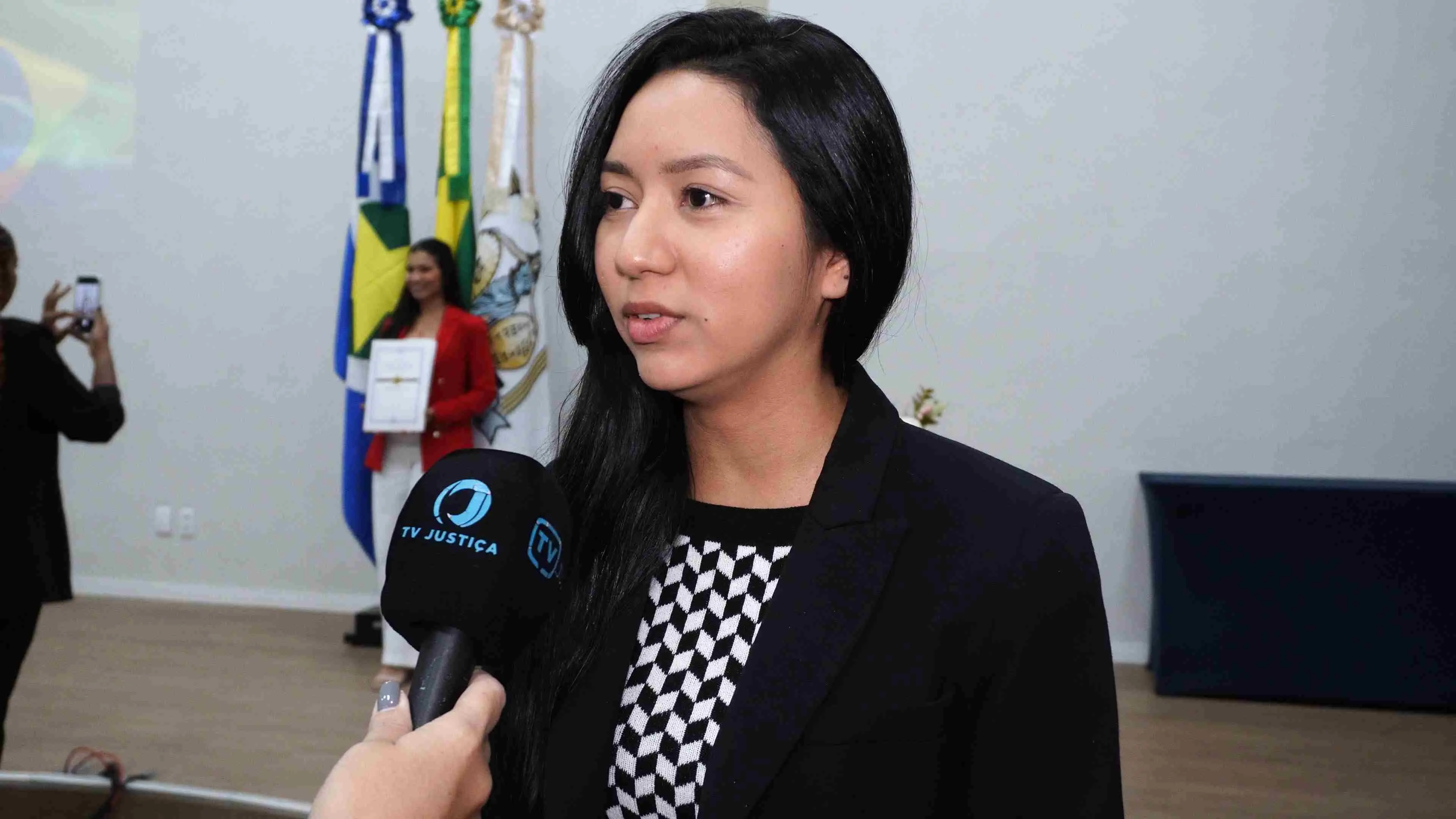 Jovem servidora concede entrevista à TV Justiça. Ela veste blazer preto e blusa com estampa geométrica. Ao fundo é possível ver parte do espaço do evento e bandeiras institucionais do Estado.