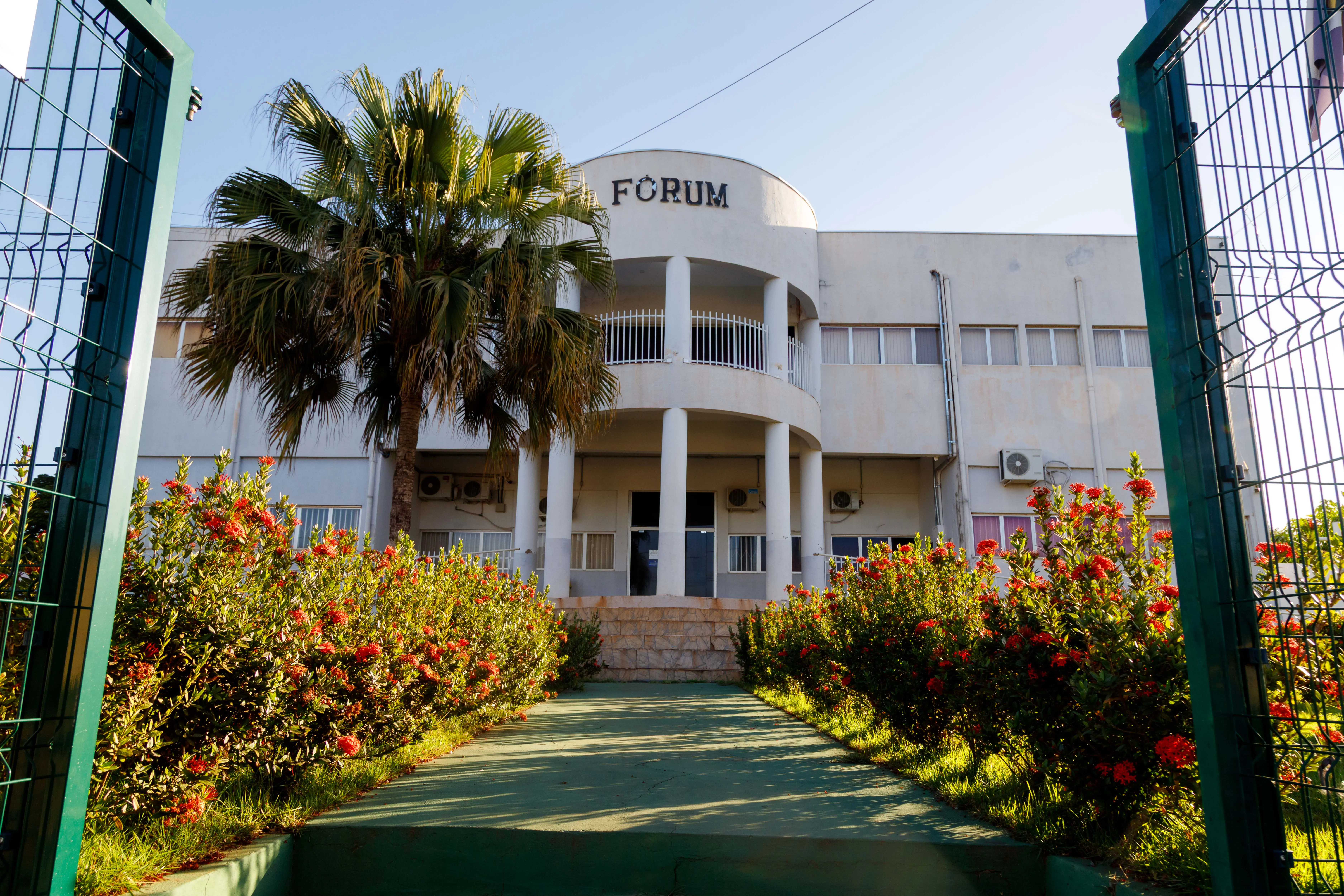  Fachada do Fórum de Juscimeira. Um prédio de dois pisos com uma palmeira, grama e flores na frente. 