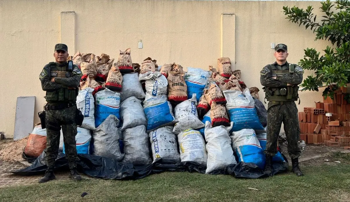 A foto mostra dois policiais militares do Juvam em pé, um de cada lado, ao lado de uma grande pilha de sacos de carvão. A pilha está em um gramado, com um muro branco e um portão à esquerda no fundo. O céu está azul e com poucas nuvens.