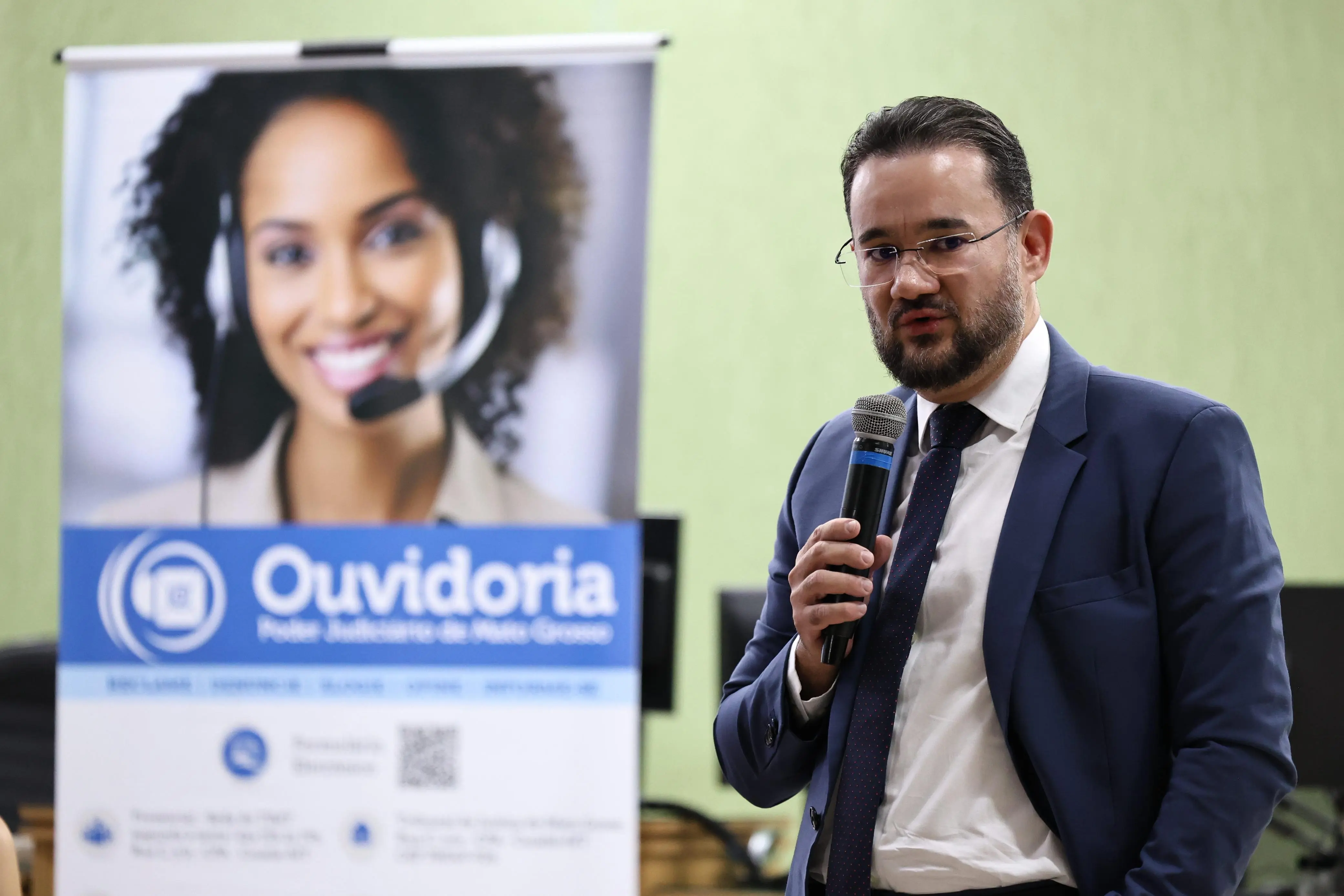 Juiz auxiliar da Ouvidoria, Bruno D'Oliveira Marques, fala ao microfone,, em pé. ele é um homem branco, de cabelo e barba castanho escuro, usando camisa branca, paletó e gravata azuis. Atrás dele, há um banner da Ouvidoria do Poder Judiciário.