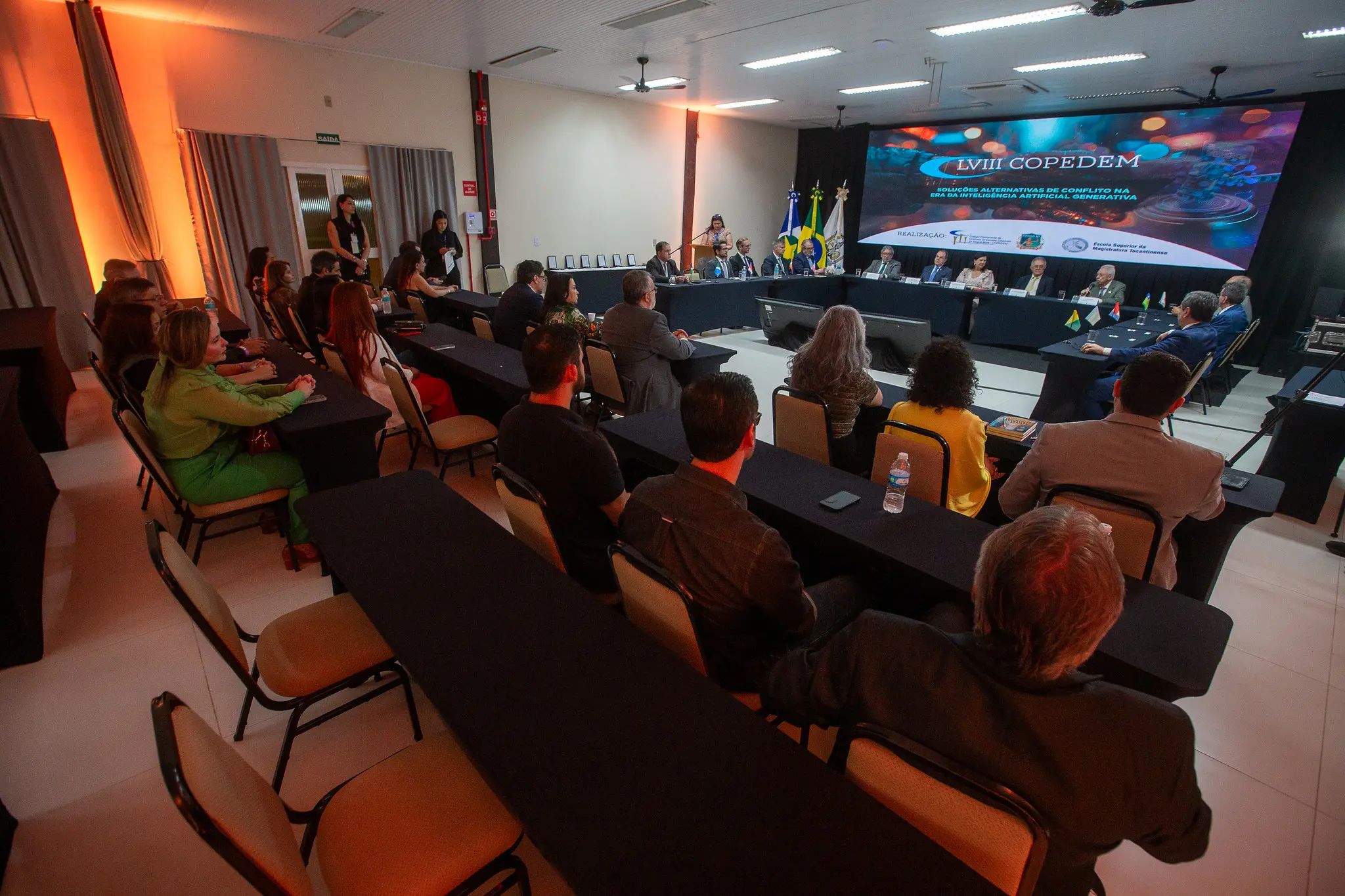 Foto em plano aberto que mostra uma sala de convenções com os participantes sentados e, à frente, a mesa de autoridades composta por diretores de escolas judiciais e demais autoridades.