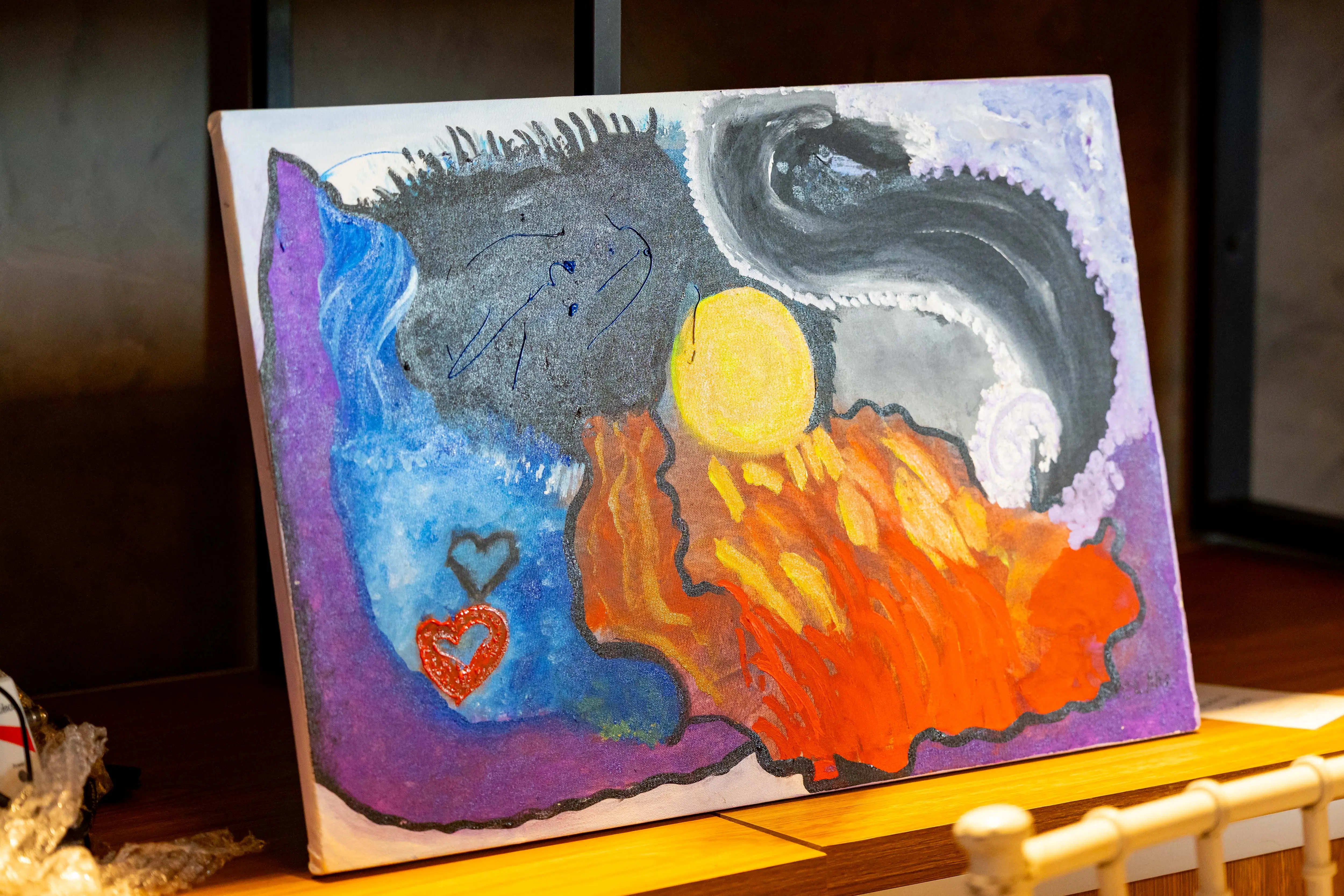 Quadro artístico exposto no estande, com sol amarelo, tons de azul, roxo e laranja, pinceladas abstratas e corações, apoiado em prateleira de madeira