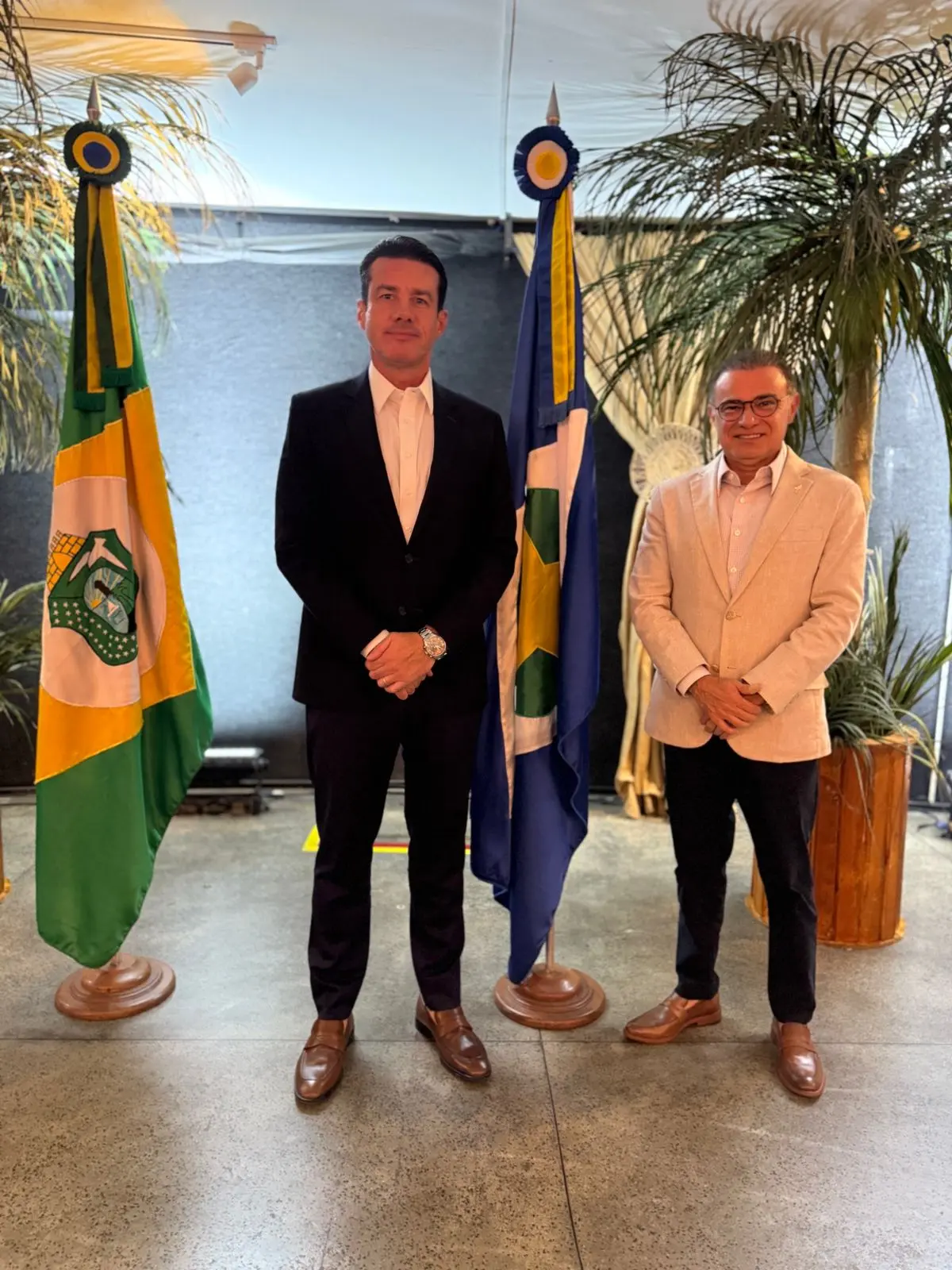 Desembargadores do TJMT e do TJ Amazonas, respectivamente, Rodrigo Curvo e Abraham Peixoto, posam para a foto sorrindo. Entre eles, há mastros com as bandeiras do Amazonas e de Mato Grosso.