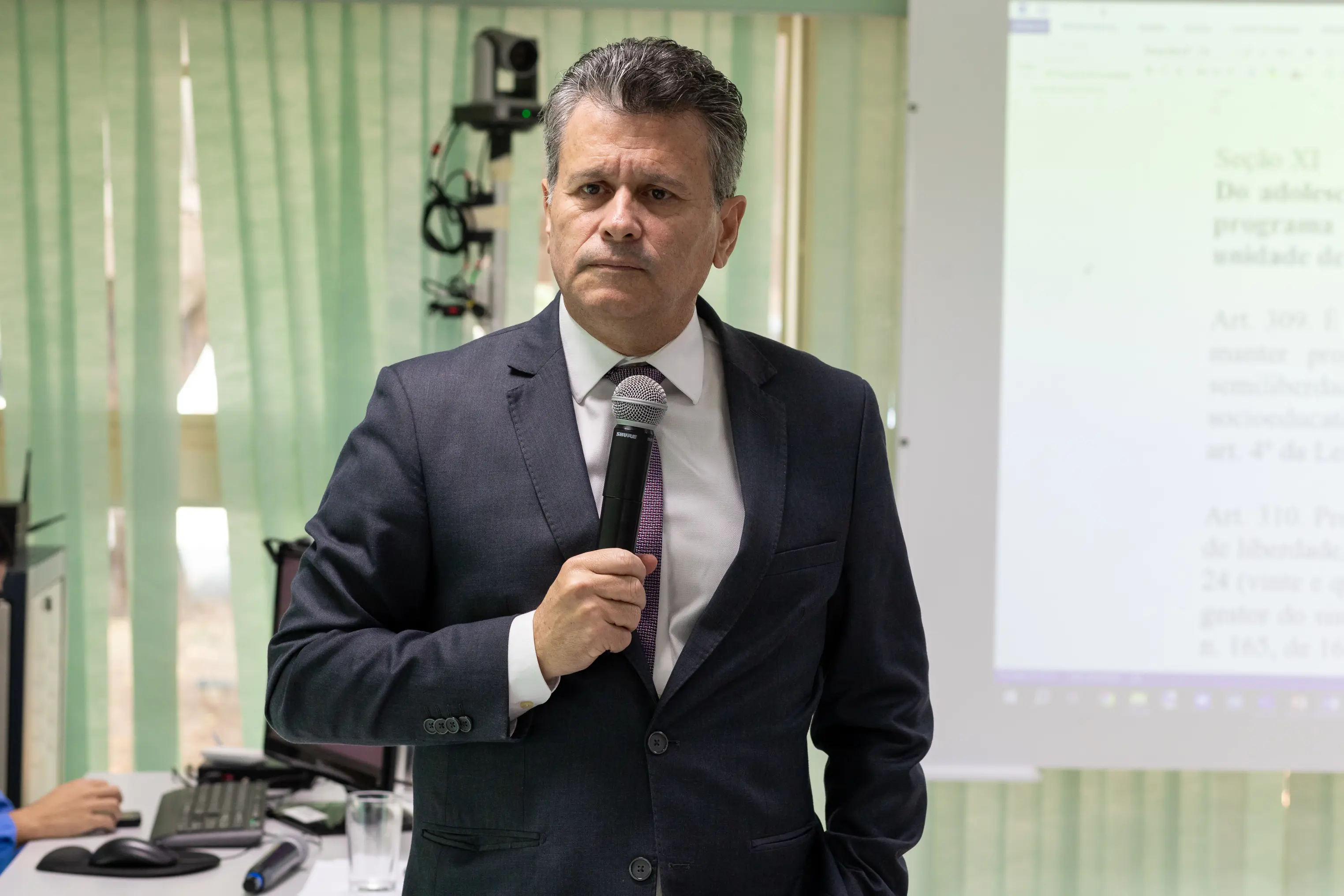 Desembargador Jones Gattass tem cabelos curtos grisalhos, expressão séria, usa terno escuro, camisa clara e gravata roxa. Segura um microfone em sala iluminada, com cortinas verdes e equipamentos ao fundo, como computador e câmera.