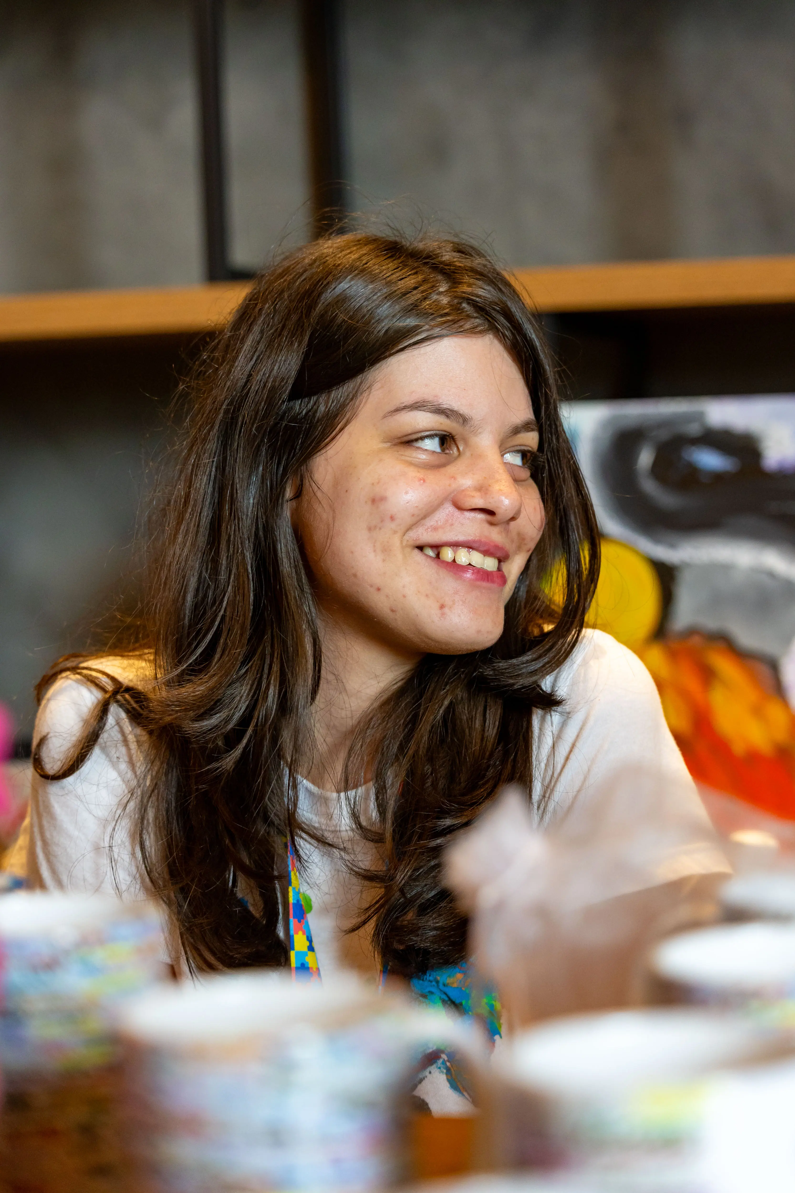 Retrato da jovem artista autista sorrindo em seu estande no TJMT Inc lusivo, com cabelos longos soltos, camiseta branca e pinturas coloridas expostas ao fundo.