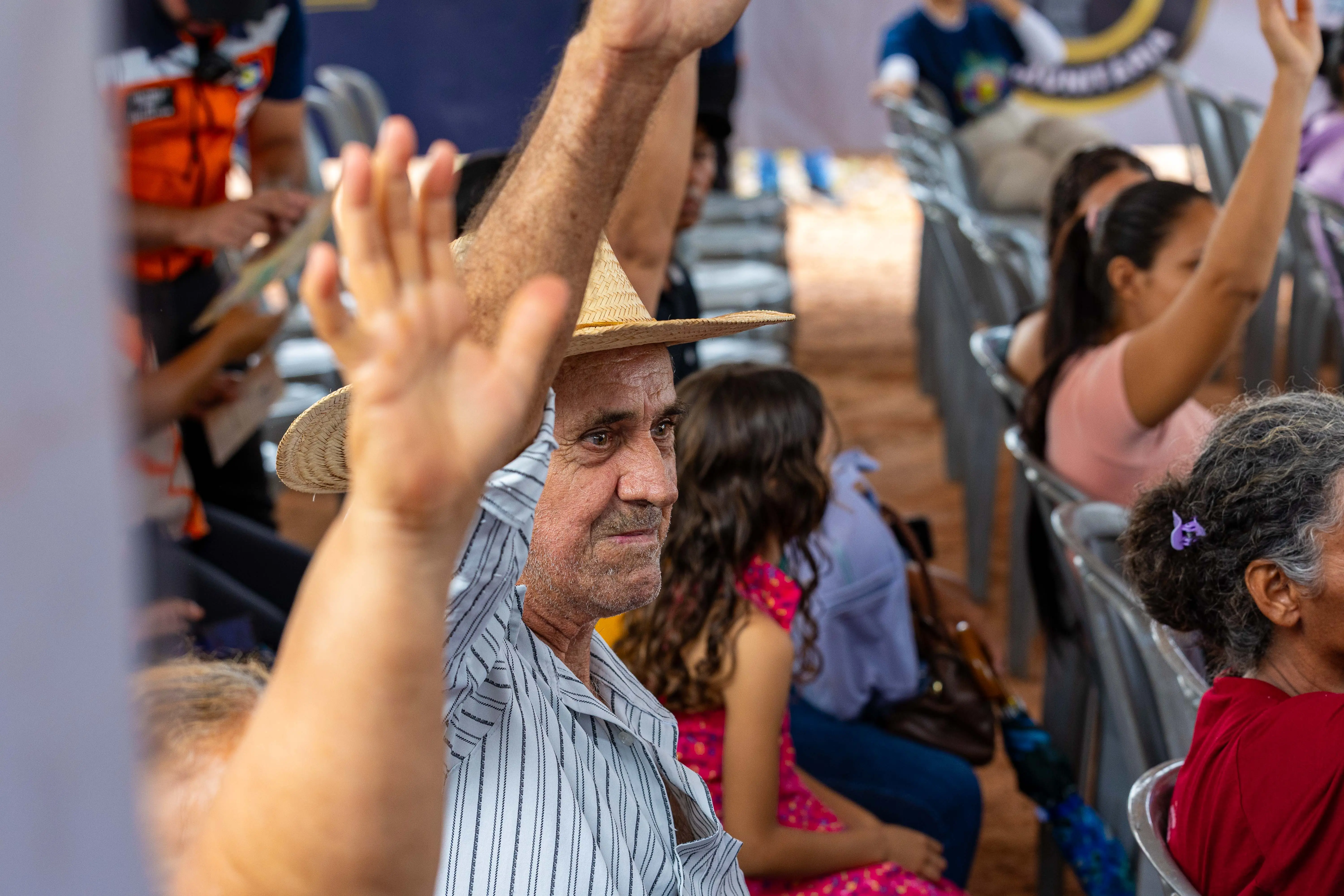  Close-up de um homem de chapéu de palha com o braço levantado em uma reunião. Ele usa uma camisa listrada. Há outras pessoas sentadas na plateia ao redor.