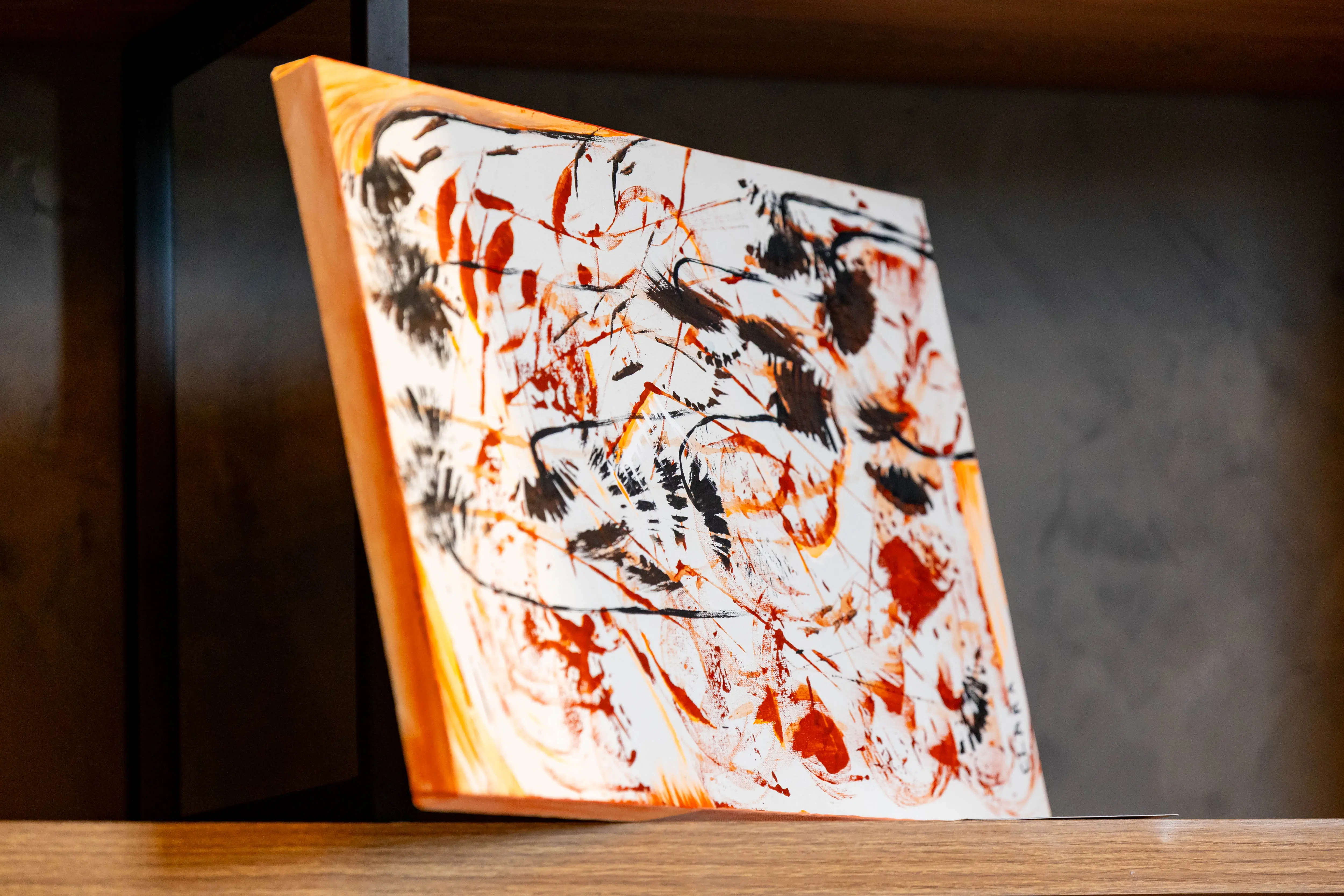 Pintura abstrata em tons de preto, laranja e vermelho, com traços marcantes e pinceladas intensas, apresentada inclinada em prateleira de madeira no estande do TJMT Inclusivo.