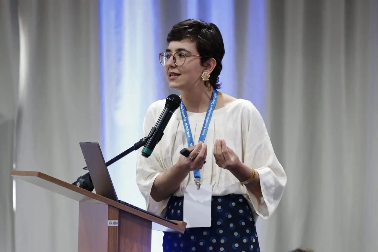 Pesquisadora da FGV, Danieli Rocha, profere palestra, falando do púlpito, em pé. Ela é uma mulhr jovem, branca, magra, usando blusa branca de mangas compridas, saia azul estampada e óculos de grau.