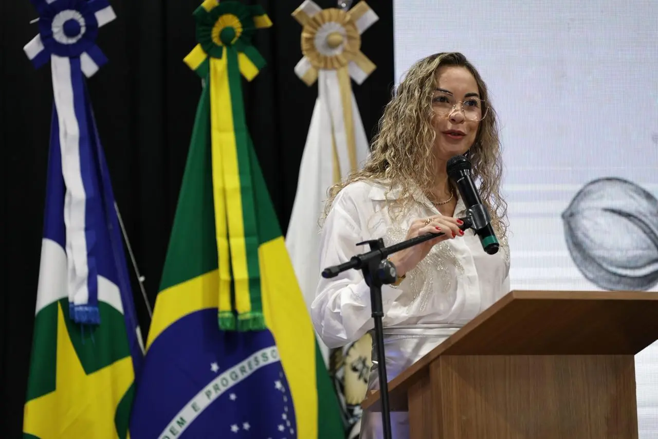 Juíza Cristiane Padim falando na tribuna. Ela é uma mulher branca, alta, de cabelos cacheados e loiros, usando camisa de manga longa branca. Atrás dela, há mastros com bandeiras do Brasil, de Mato Grosso e do Poder Judiciário.