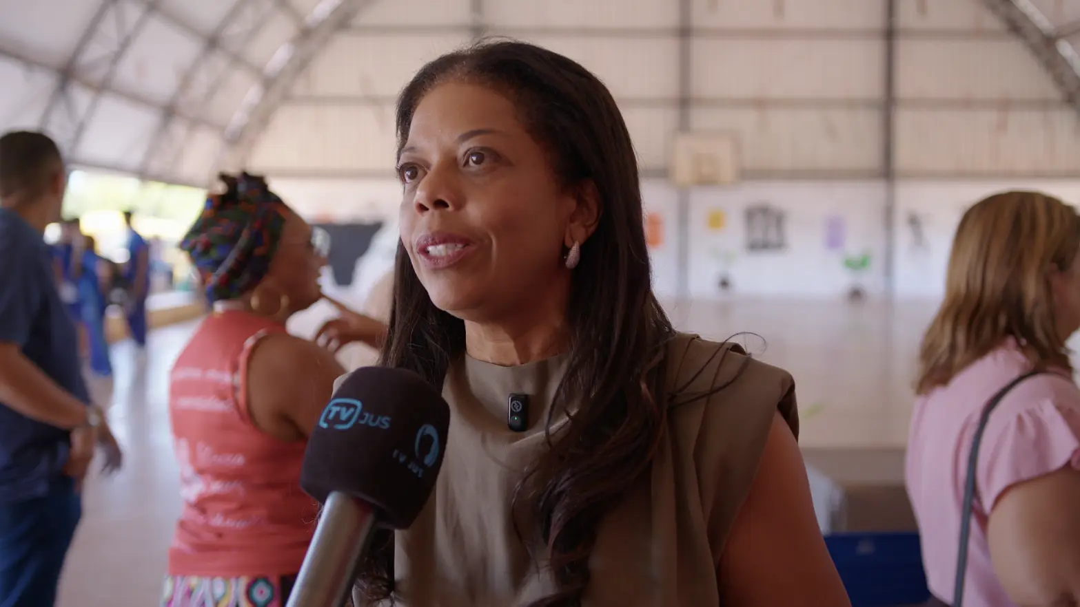 Ju�za Renata Evaristo durante entrevista � TV Justi�a. Ela � uma mulher negra, de cabelos longos e castanhos, usando roupa verde militar. ela est� no gin�sio de uma escola estadual. 