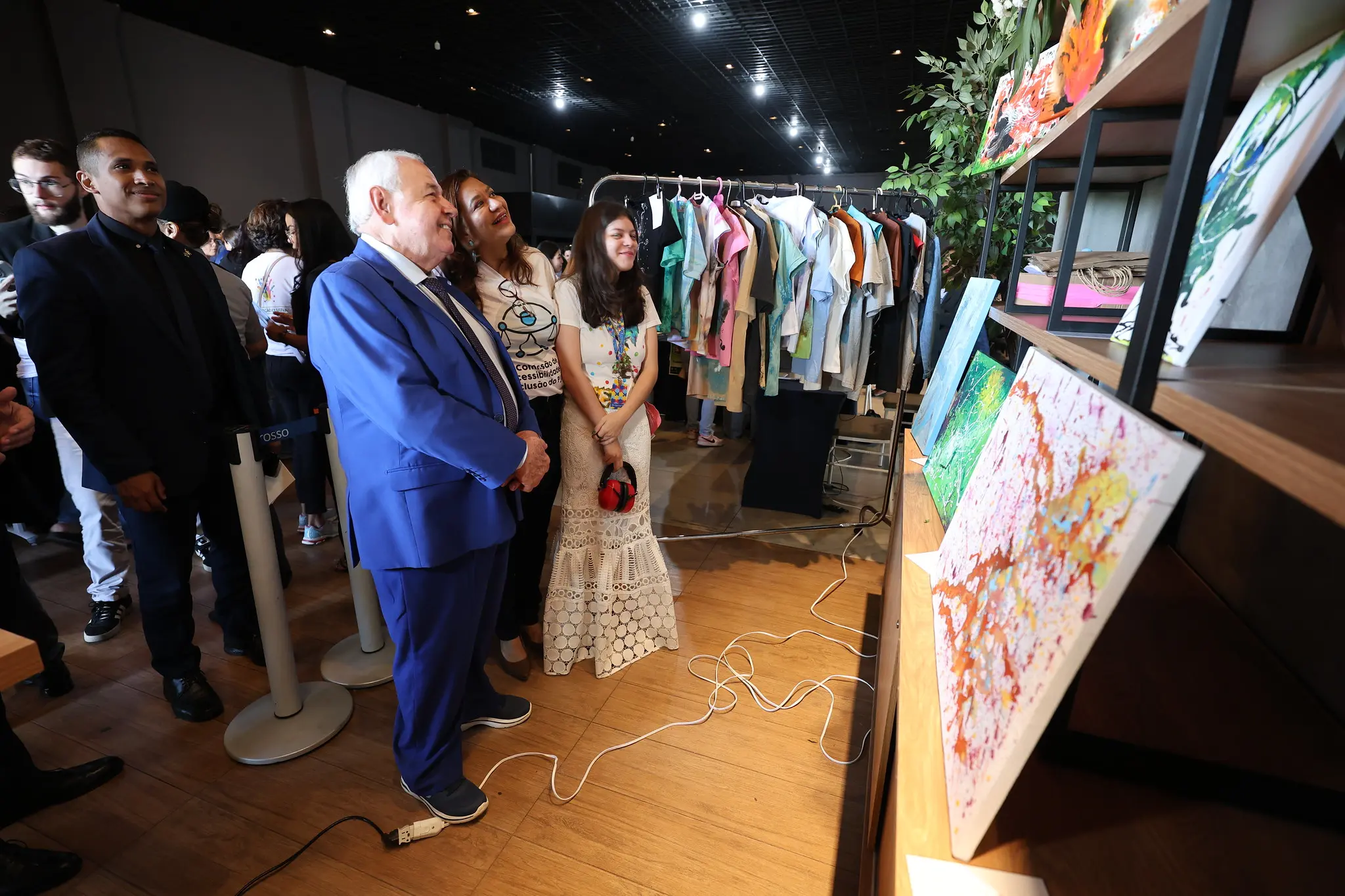 O presidente do TJMT e equipe observam quadros coloridos alinhados em prateleira no TJMT Inc lusivo, enquanto a jovem artista e familiares acompanham sorrindo a visita ao estande.