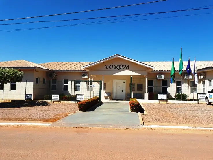 Fachada do Fórum da Comarca de Nova Monte Verde, em Mato Grosso. O prédio térreo, de cor clara, tem acesso central com rampa e a identificação “Fórum” acima da entrada. À direita, mastros exibem bandeiras institucionais. O entorno é aberto, com jardim simples e via pavimentada.
