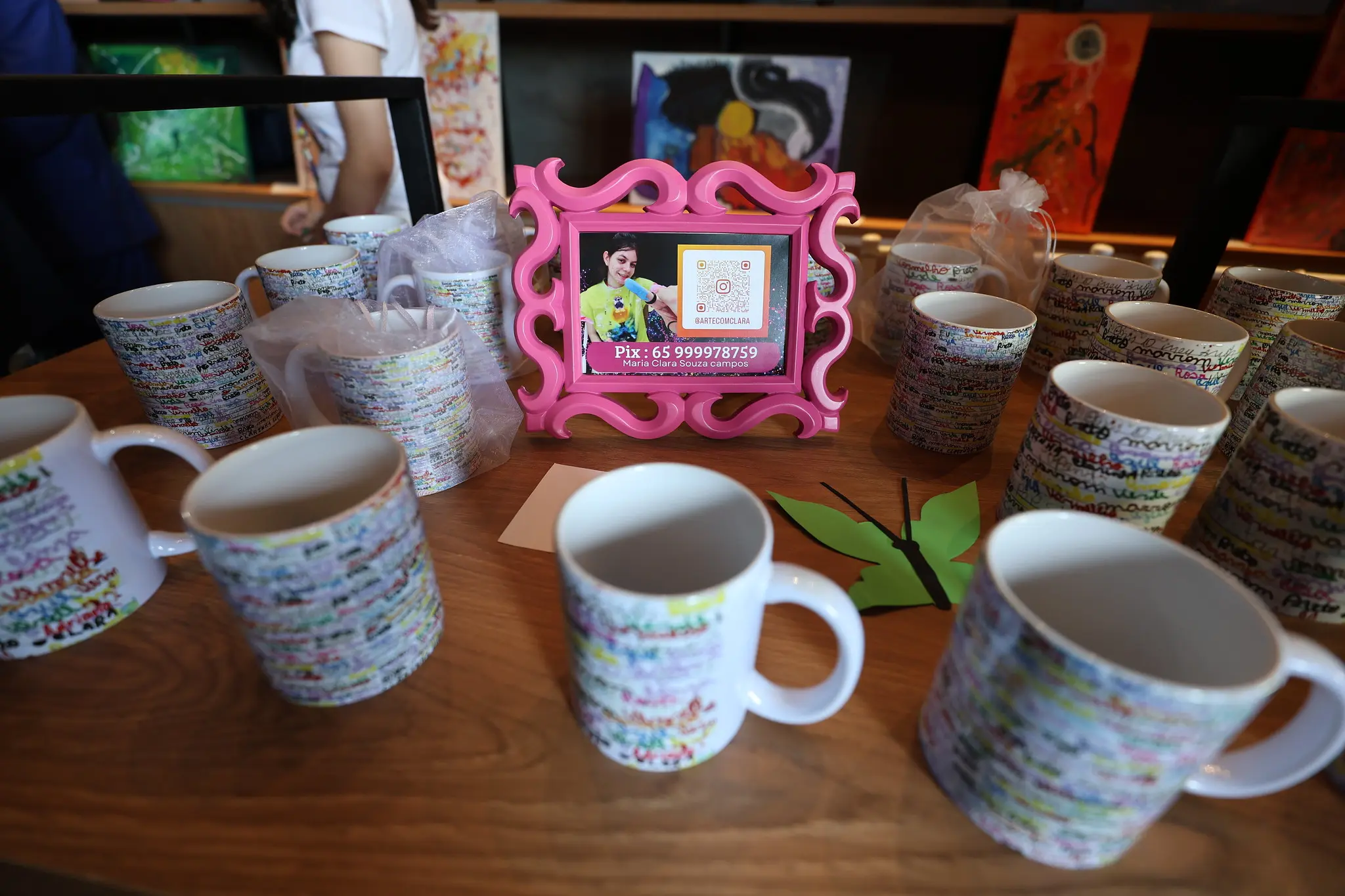 Mesa de madeira exibe canecas personalizadas e moldura rosa com foto e dados de contato da jovem artista, cercada por pessoas apreciando as peças.