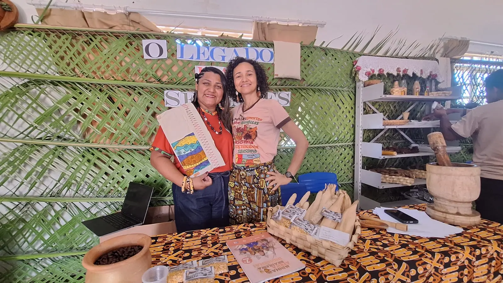 Diretora e professora da Escola Estadual Quilombola de Mata Cavalo posam para a foto sorrindo, no stand que fala sobre o projeto O Legado das Sementes.