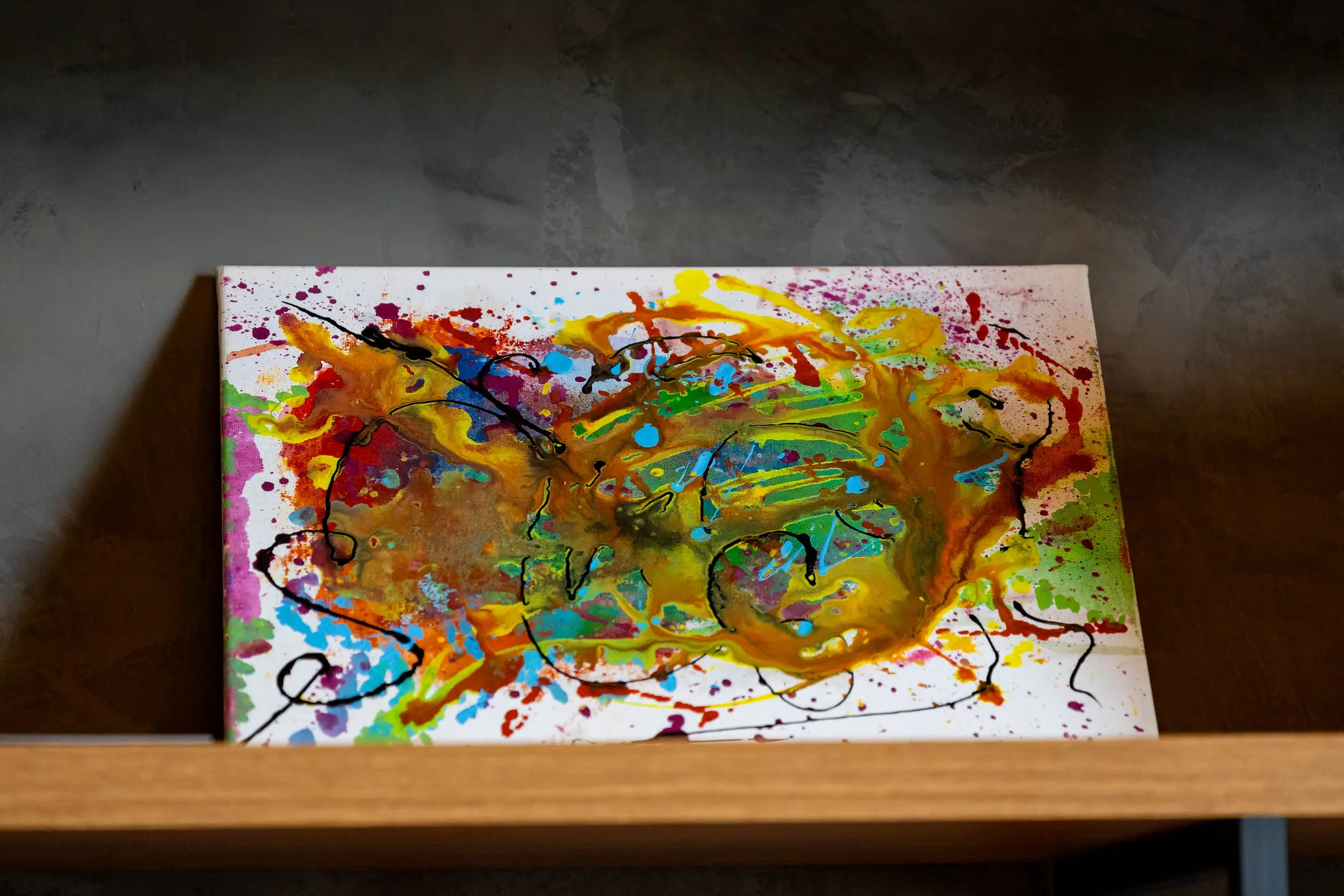 Tela abstrata repleta de cores vibrantes — amarelo, verde, rosa e azul — exposta em prateleira de madeira no estande artístico do evento TJMT Inclusivo, em ambiente interno iluminado.