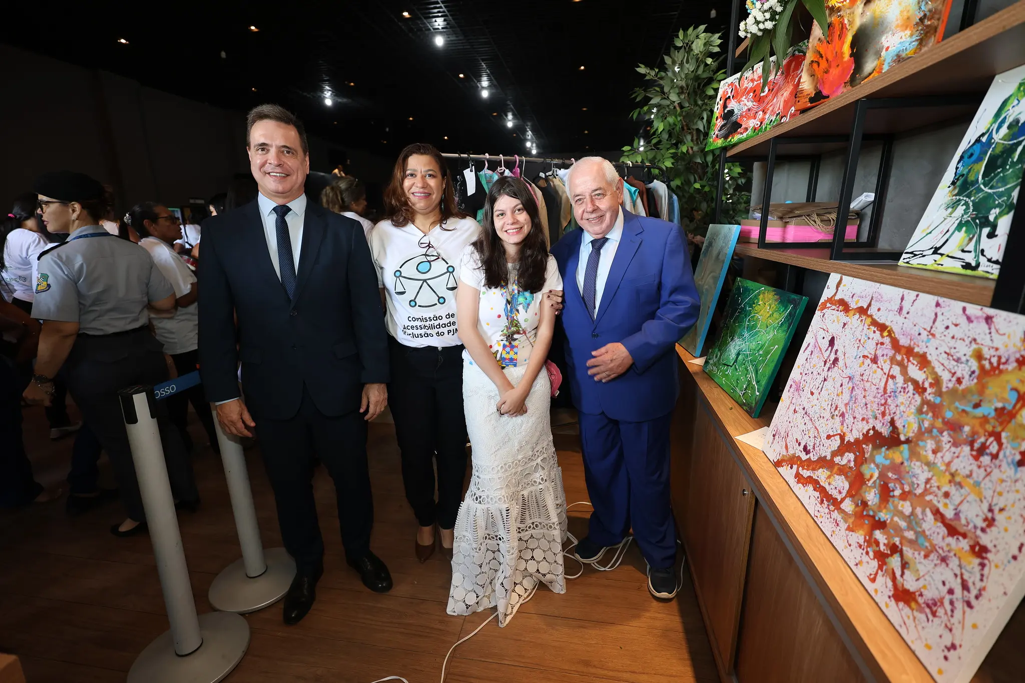 Maria Clara com o presidente do TJMT, posa sorrindo em corredor iluminado do Centro de Eventos do Pantanal, ao lado de prateleiras com quadros coloridos expostos.