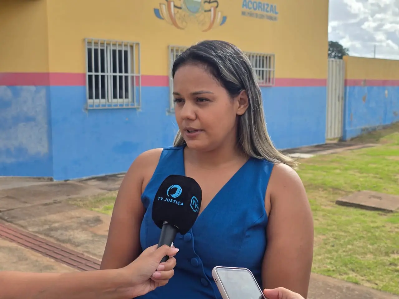 Foto horizontal em plano médio da conselheira tutelar Stefany de Arruda, durante entrevista à TV Justiça. Ela é uma mulher negra, de cabelos longos com mechas loiras, usando colete azul. Ela está em frente a um prédio público onde funciona o Centro de Convivência de Idosos de Acorizal.
