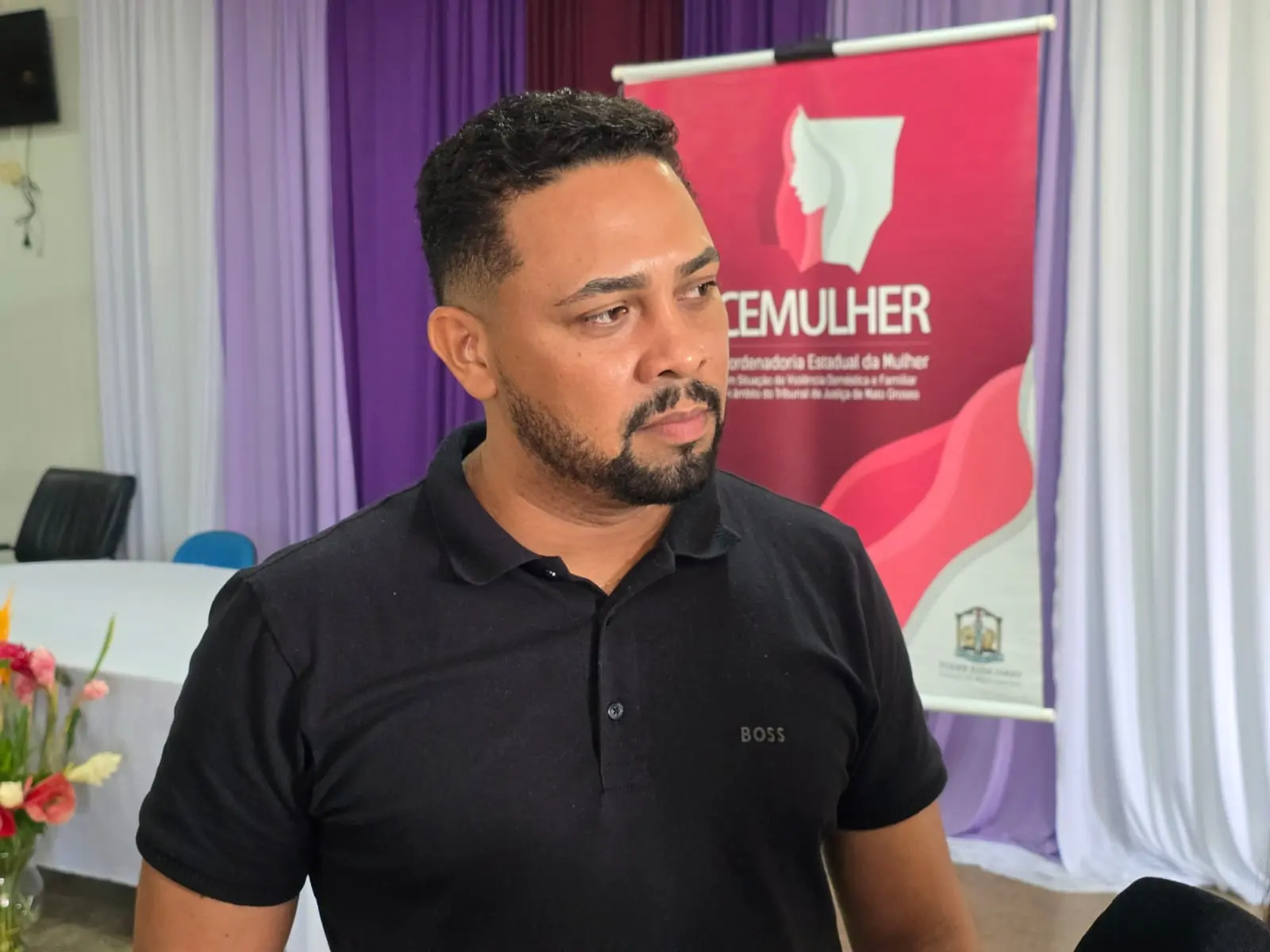 Foto horizontal em plano médio que mostra o secretário de Assistência Social de Acorizal, Roberth Lima. Ele é um homem negro, de olhos castanhos, cabelos e barba pretos, usando camiseta polo preta. Atrás dele, há banner da Cemulher. 