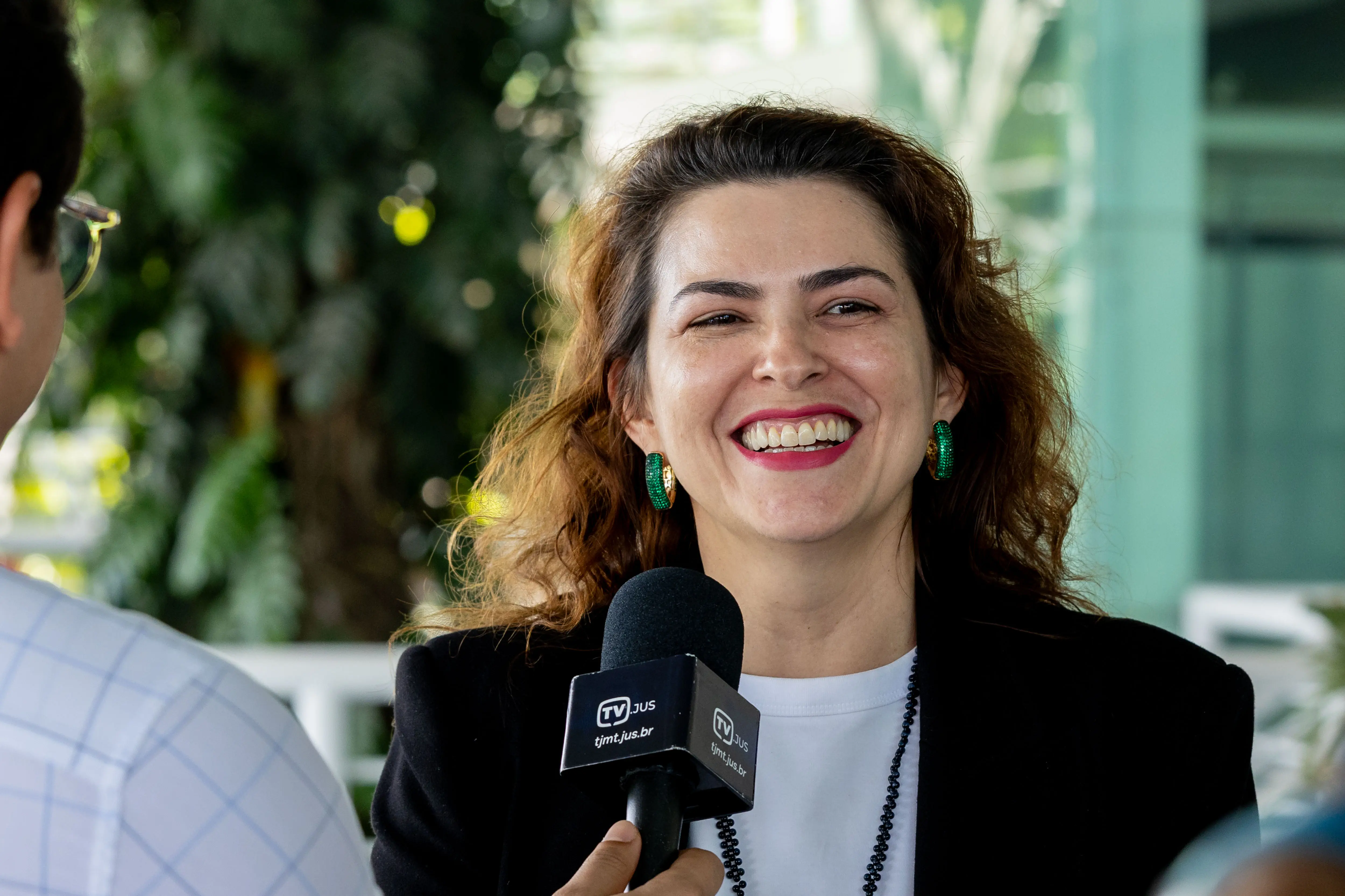 Mulher branca sorridente, com cabelos castanhos ondulados e batom rosa. Veste blazer preto e brincos verdes redondos. Ela segura um microfone com a logo 