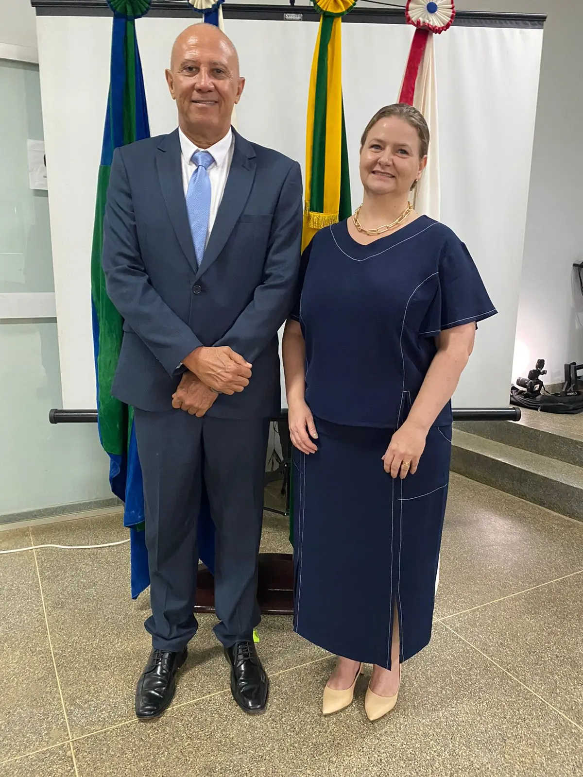 A juíza diretora do Foro da Comarca de Cotriguaçu, Gezicler Luiza Sossanovicz Artilheiro, posa ao lado de Maurício César Bento, responsável pelo Cartório do 1º Ofício de Registro de Imóveis, Títulos e Documentos do município. Ao fundo aparecem bandeiras oficiais.