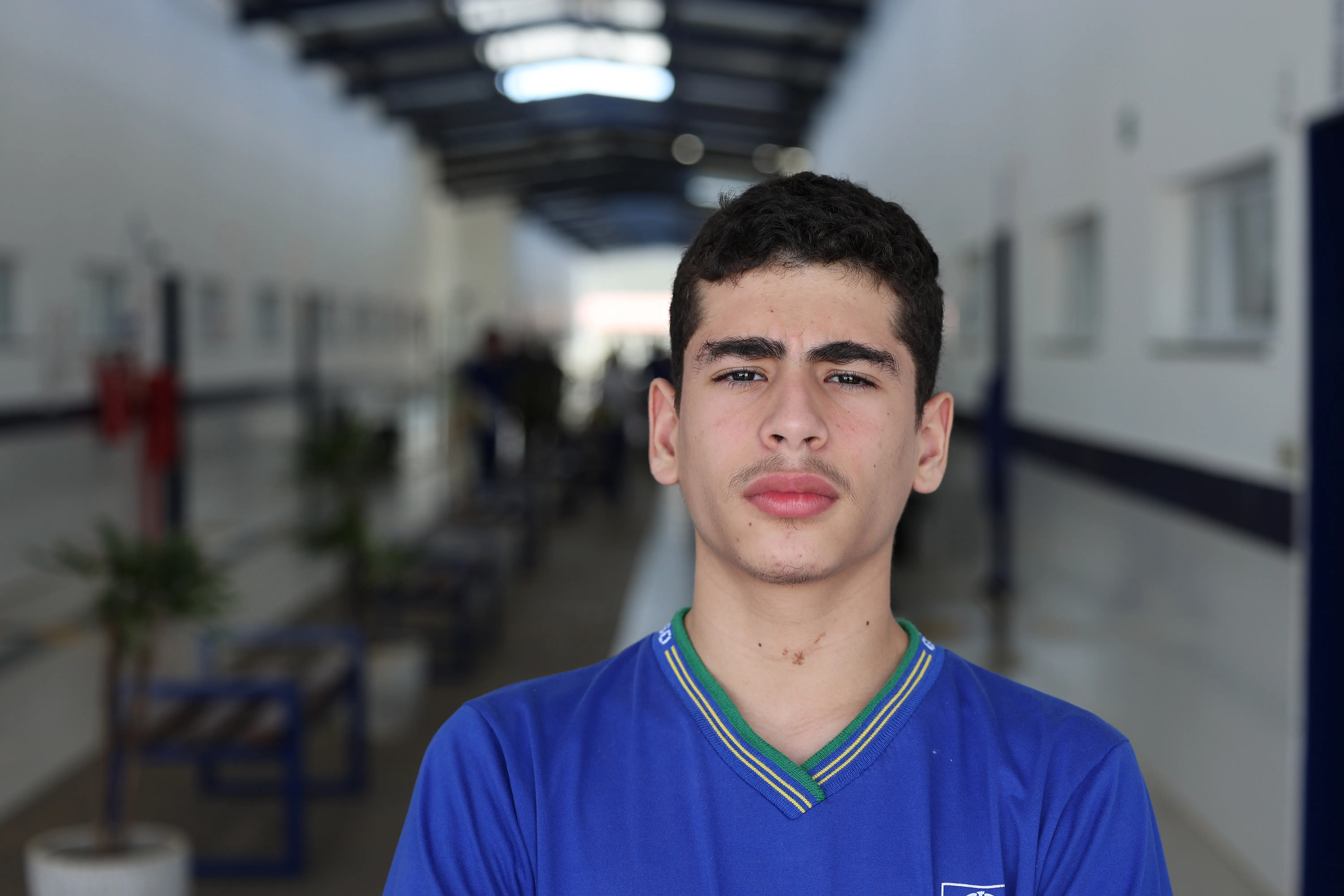 Retrato de Cauê Alves Marrafon. Jovem de pele clara e cabelos curtos e escuros. Ele mantém uma expressão séria e direta para a câmera. Veste o uniforme escolar azul. O cenário de fundo é o corredor interno da instituição.