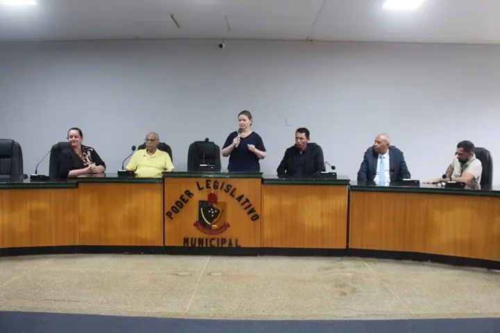 Juíza fala ao microfone na mesa do plenário da Câmara Municipal de Cotriguaçu. Ao lado dela estão autoridades sentadas. Na frente da mesa aparece o brasão do Poder Legislativo Municipal