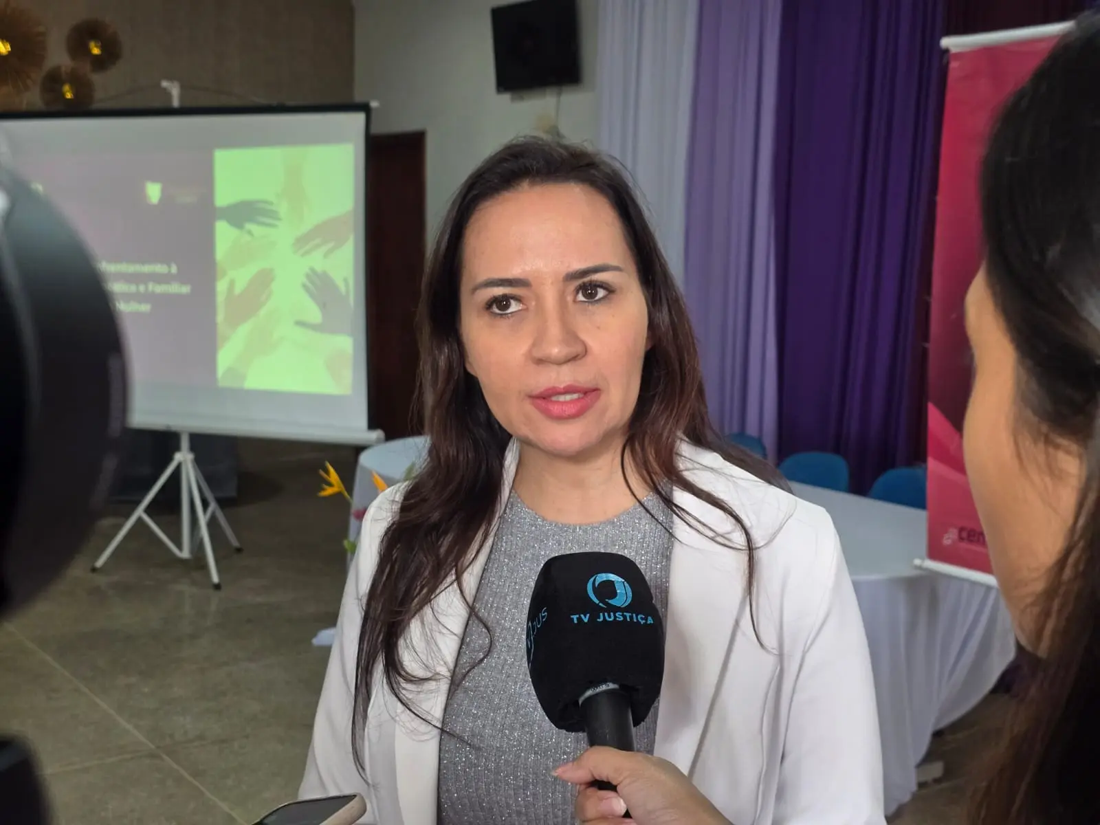 Foto horizontal em plano médio que mostra a juíza Tatyana Lopes, durante entrevista à TV Justiça. Ela é uma mulher parda, de cabelos longos, lisos e castanhos, olhos castanhos, usando blusa prateada e blazer branco. Ela está em um salão onde realizou palestra da Cemulher.