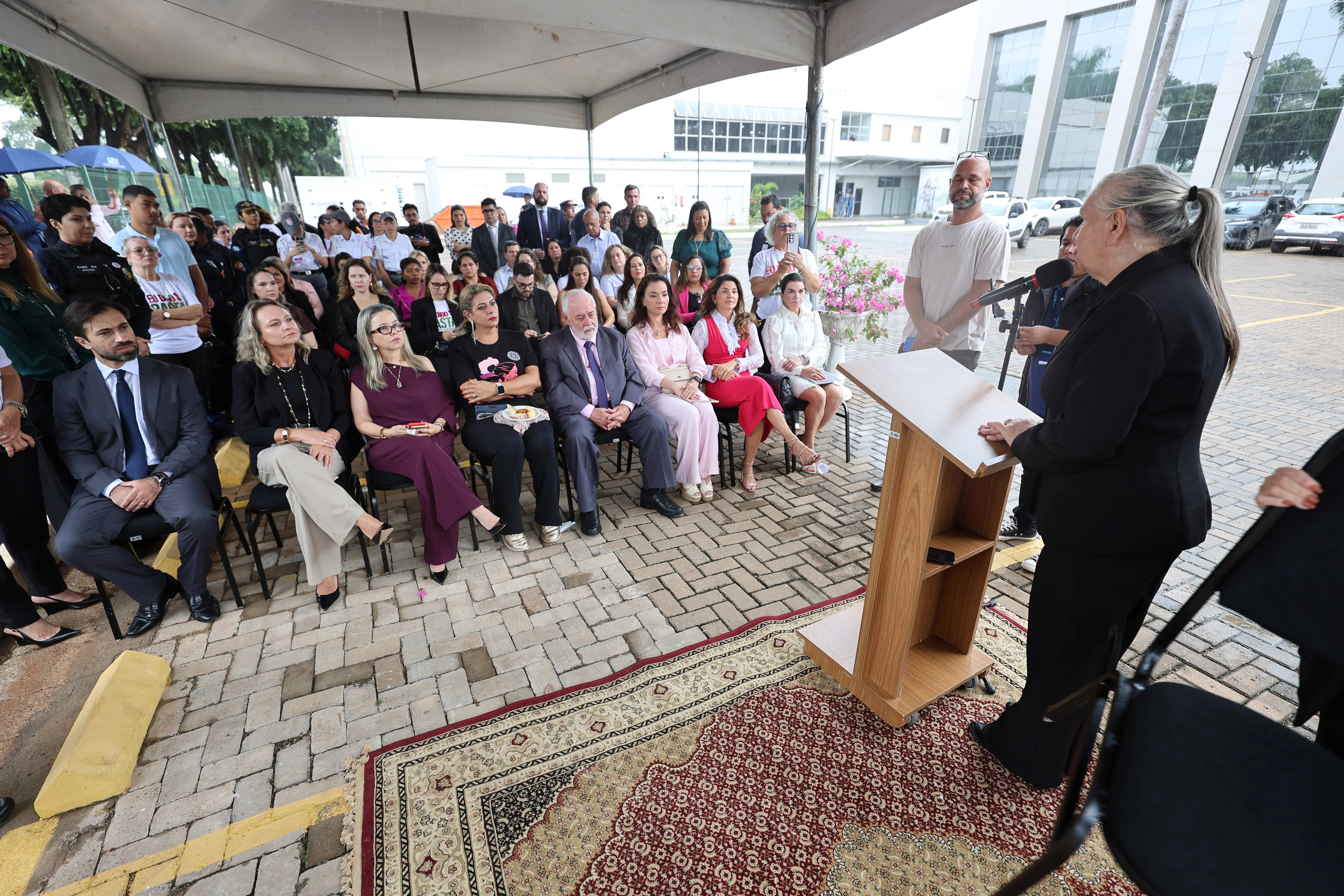  O Gemini disse Sob uma tenda, uma mulher discursa em um púlpito para uma plateia sentada e atenta. O grupo é diversificado, composto por homens e mulheres em trajes formais. O evento ocorre em uma área externa pavimentada, com prédios ao fundo.