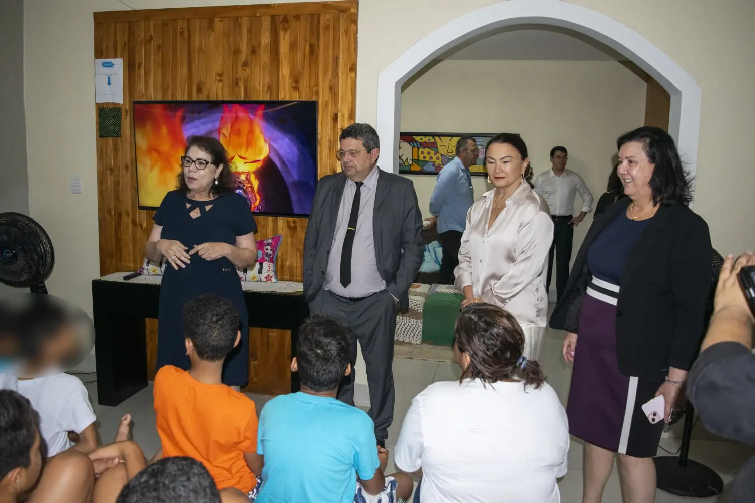 A imagem mostra a comitiva do Judiciário em pé, conversando com as crianças. Elas estão sentadas e olham para os magistrados.