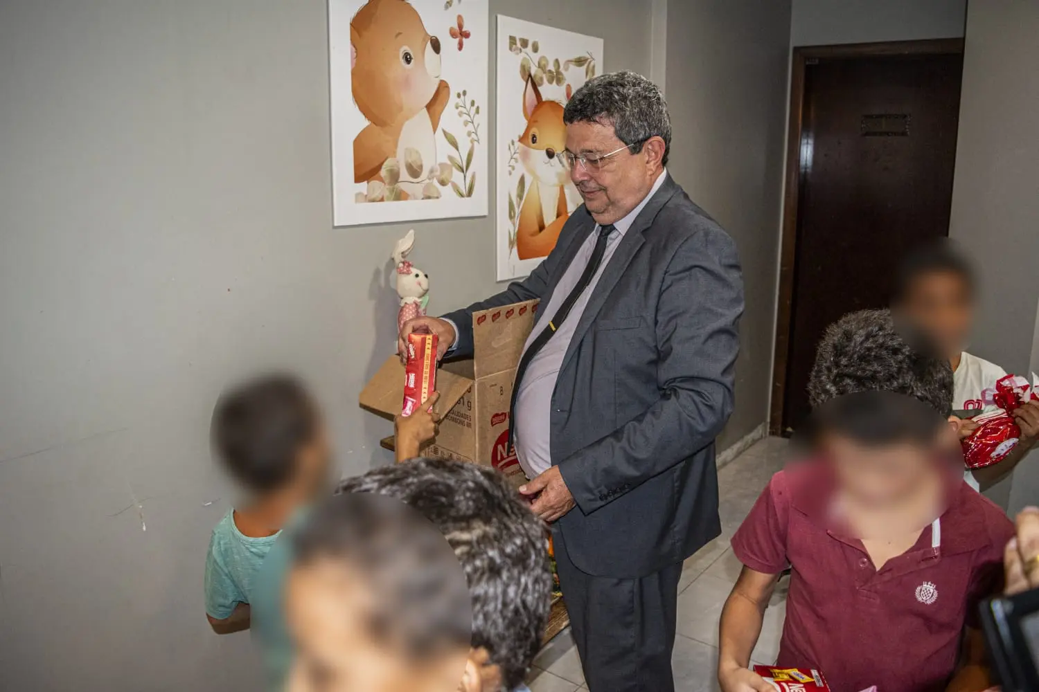 Corregedor está em pé, rodeado por crianças. Ele segura uma caixa e distribui chocolates.