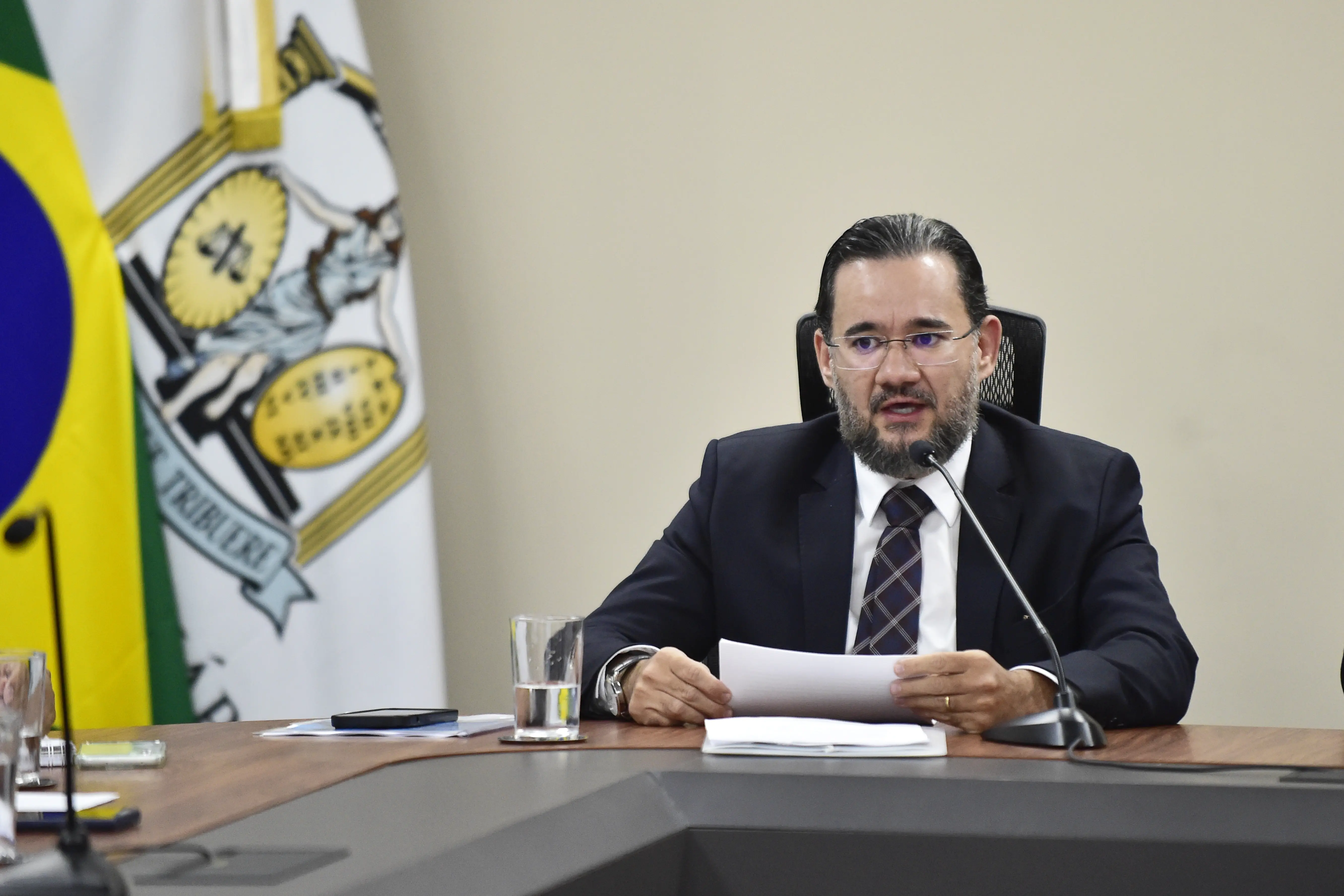 Juiz Bruno D’Oliveira sentado em torno de uma mesa de reuniões e falando ao microfone. Ele é um homem branco, de cabelo e barba grisalhos, usando óculos de grau, camisa branca, gravata e paletó preto. No canto esquerdo da foto, há dois mastros com as bandeiras do Brasil e do Poder Judiciário. 