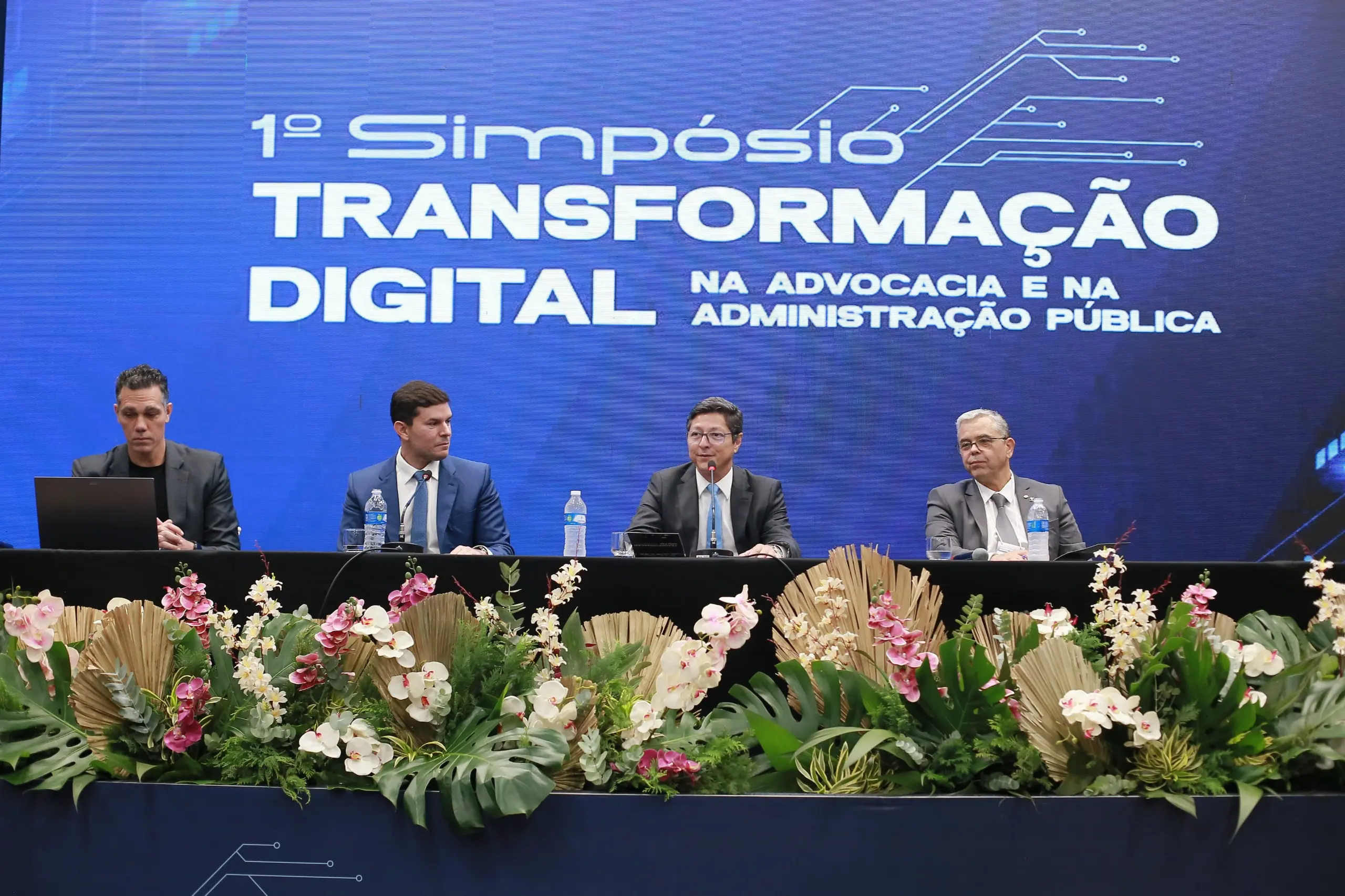 palestrantes, Maximiliano Carvalho e Alisson Alencar, o desembargador Lídio Modesto e o juiz Hildebrando Costa Marques. Todos são homens brancos, usando terno e gravata. Atrás deles há um grande telão com a logo do 1º Simpósio de Transformação Digital. Na frente da mesa, há flores e folhas.